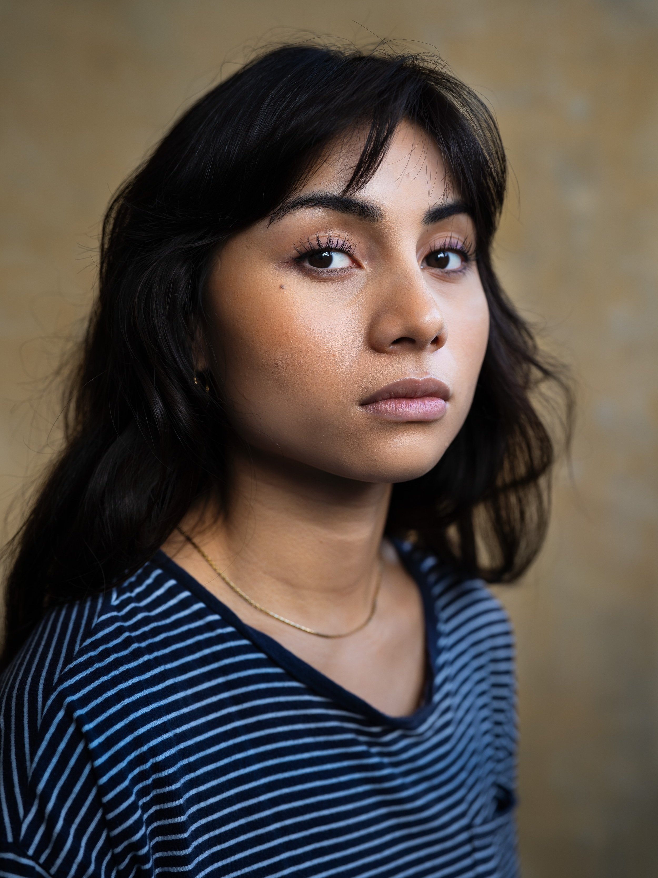 Portrait of a woman with long dark hair wearing a striped shirt and a subtle necklace, looking into the distance against a plain background.