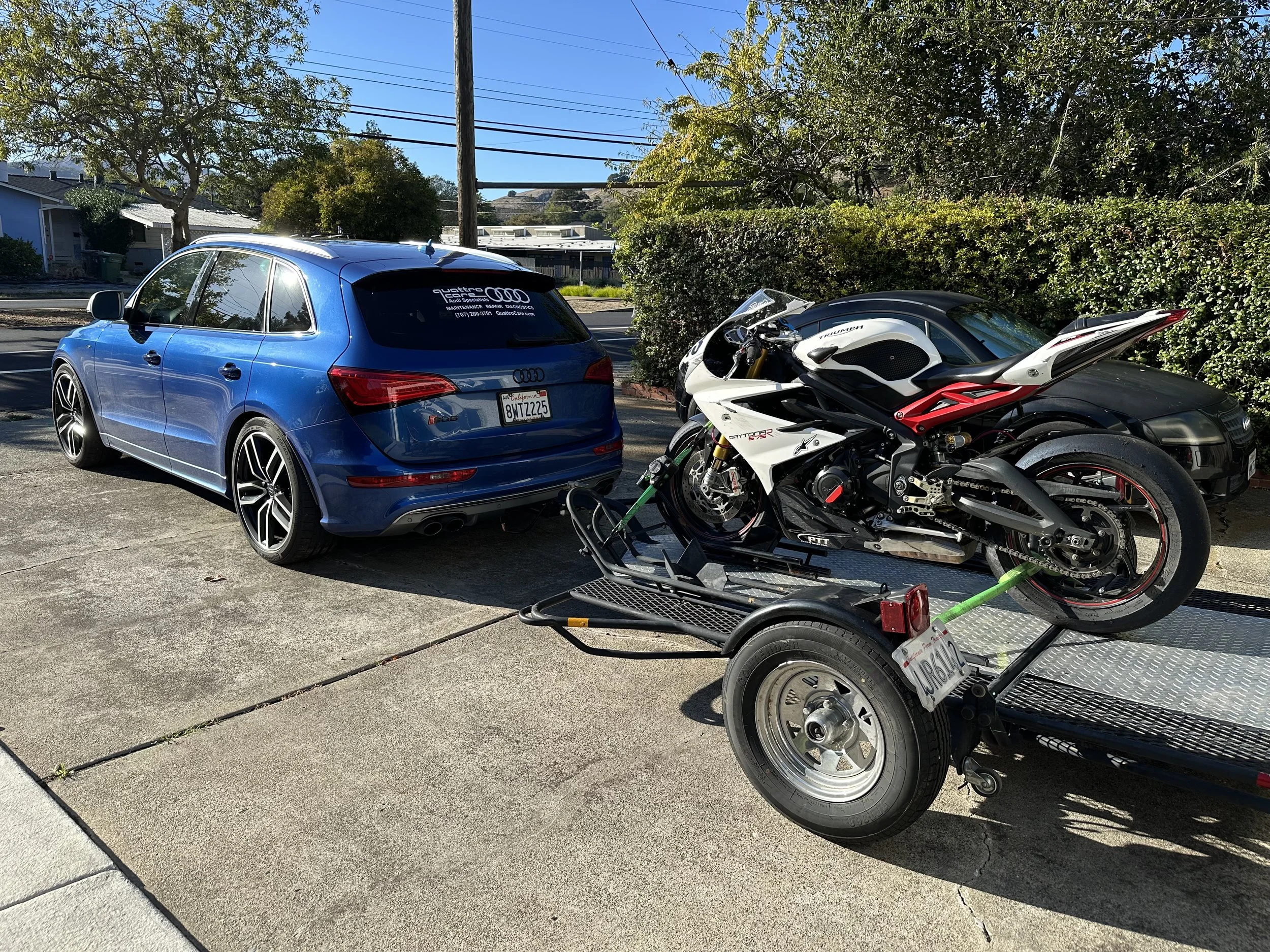 A blue Audi SQ5 parked on a driveway, with a white and black motorcycle on a trailer attached to the back of the car. The trailer has a license plate, and the scene is set in a residential area with bushes, trees, and houses in the background.