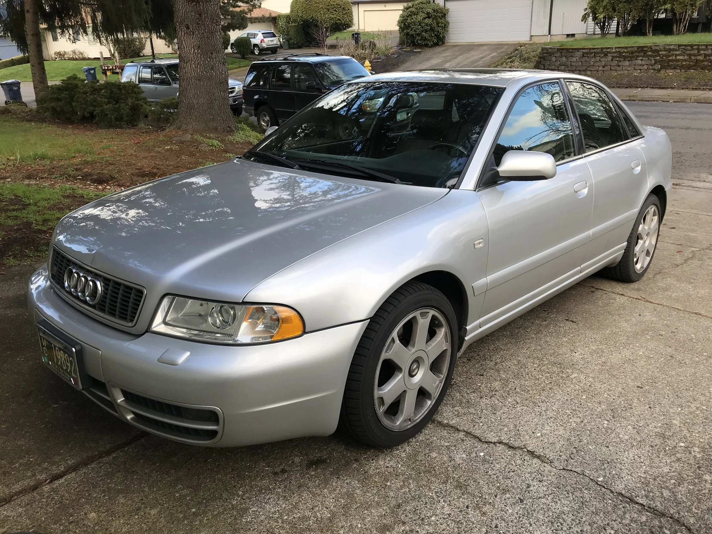 Silver Audi S4 sedan parked on a concrete driveway in a residential neighborhood, with trees, grass, and other cars in the background.
