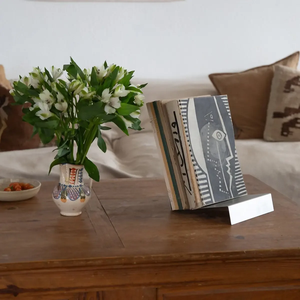 A wooden table with a small vase of white flowers, a bowl of snacks, and a book holder displaying a book or magazine with a face illustration.