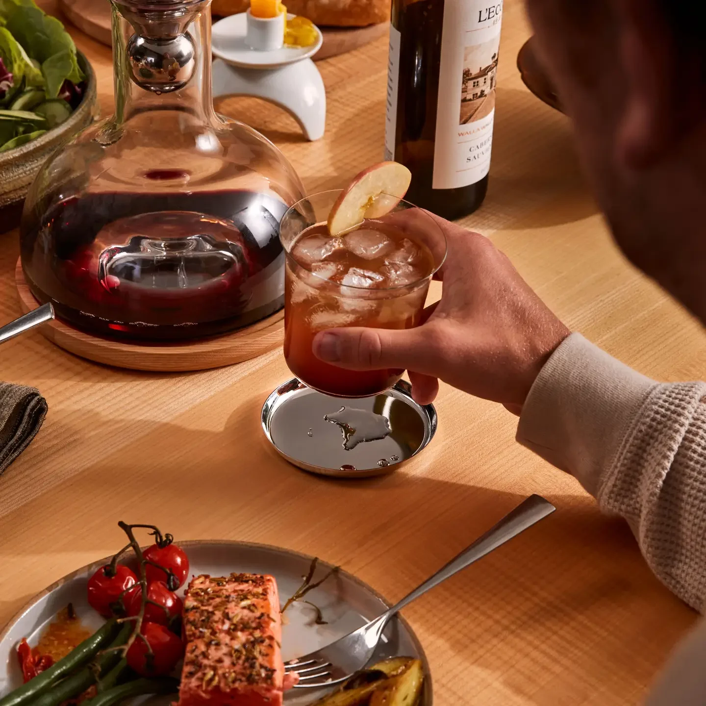 A person holding a glass of iced tea with a lemon slice, next to a decanter of red wine on a wooden table, with a plate of food, grapes, and candles nearby.