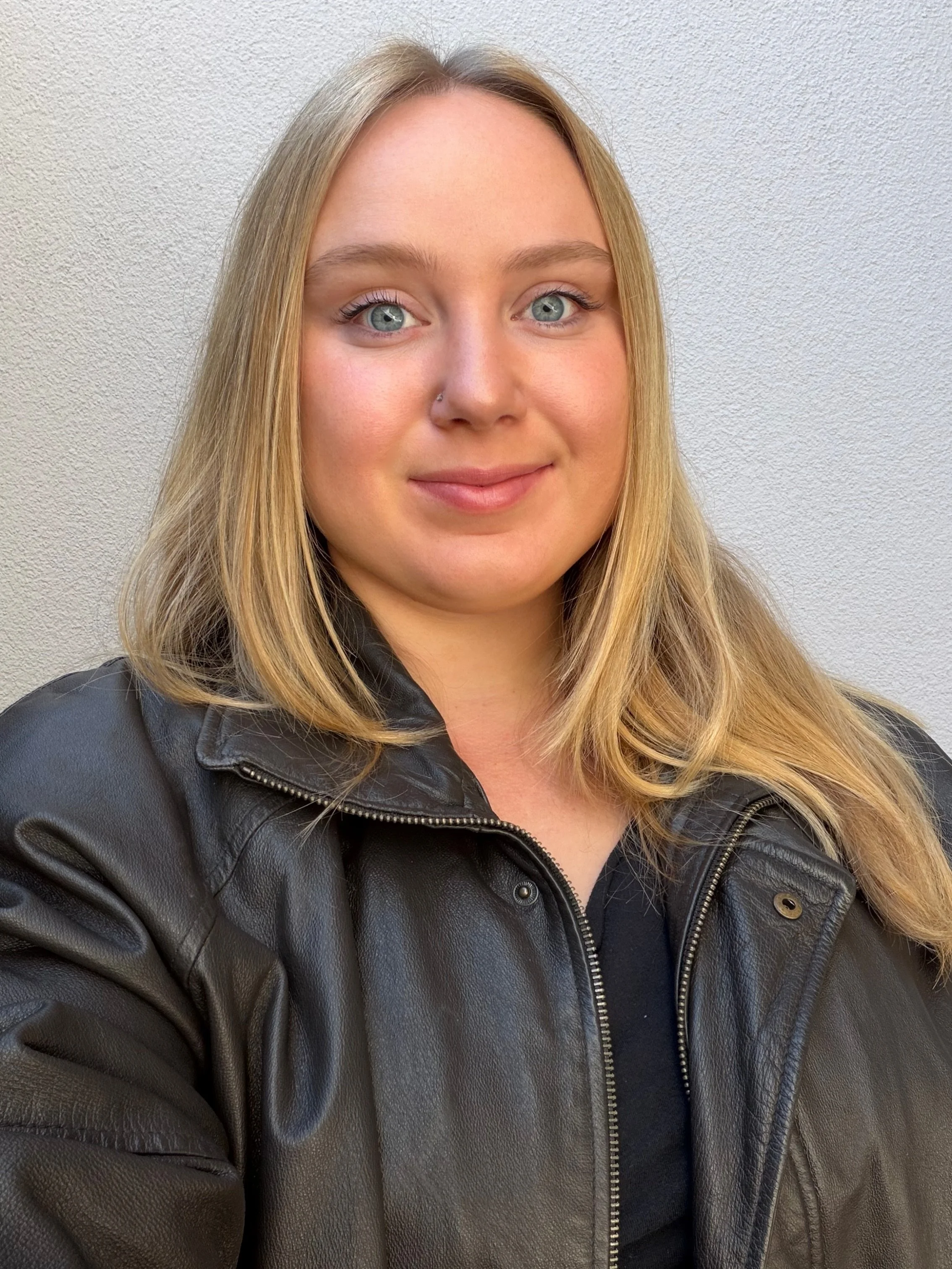 A young woman with blond hair, blue eyes, and fair skin wearing a black leather jacket standing in front of a plain, light-colored wall.