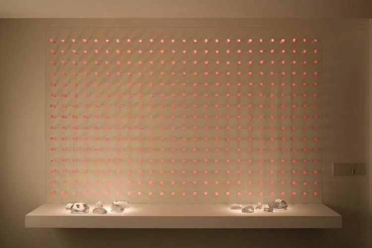 luz ou cor?
De início parecem apenas objetos pintados de uma cor opaca, espécie de batons arranjados sobre um painel vertical.
Nem parece que aquelas cores, um vermelho terroso, um azul cinzento, são, na verdade, luz. Apenas quando o ambiente se