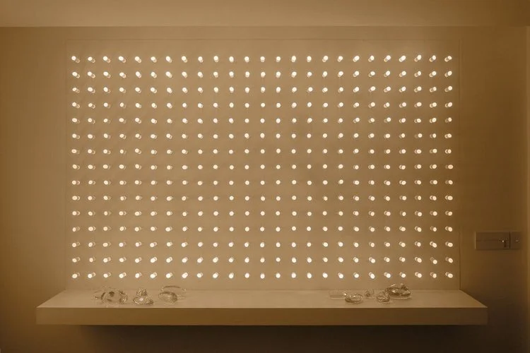 luz ou cor?
De início parecem apenas objetos pintados de uma cor opaca, espécie de batons arranjados sobre um painel vertical.
Nem parece que aquelas cores, um vermelho terroso, um azul cinzento, são, na verdade, luz. Apenas quando o ambiente se