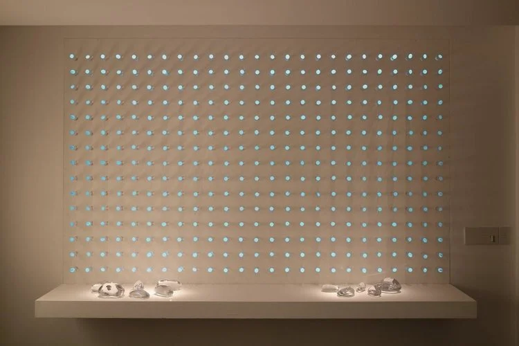 luz ou cor?
De início parecem apenas objetos pintados de uma cor opaca, espécie de batons arranjados sobre um painel vertical.
Nem parece que aquelas cores, um vermelho terroso, um azul cinzento, são, na verdade, luz. Apenas quando o ambiente se