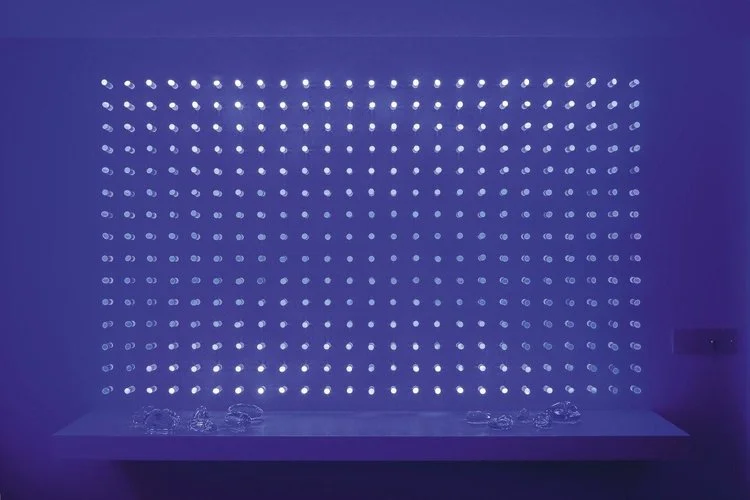 luz ou cor?
De início parecem apenas objetos pintados de uma cor opaca, espécie de batons arranjados sobre um painel vertical.
Nem parece que aquelas cores, um vermelho terroso, um azul cinzento, são, na verdade, luz. Apenas quando o ambiente se