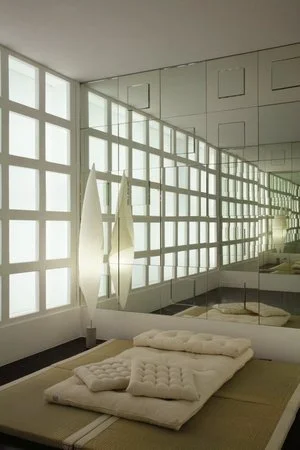 Poderíamos estar num quarto japonês — e estamos: um tatame, delicadas luminárias de papel microplissado, e uma divisória que quase se faz passar por aquelas de papel arroz emolduradas em quadriculado. Atrás da divisória, a luz difusa, etérea, como co