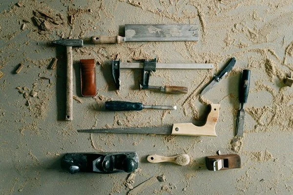Collection of woodworking tools including a saw, chisel, hammer, and hand plane on a rough wooden surface.
