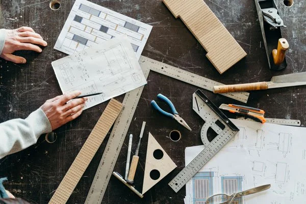 Architectural blueprints and drafting tools on a dark workspace, with hands holding a pen and ruler.