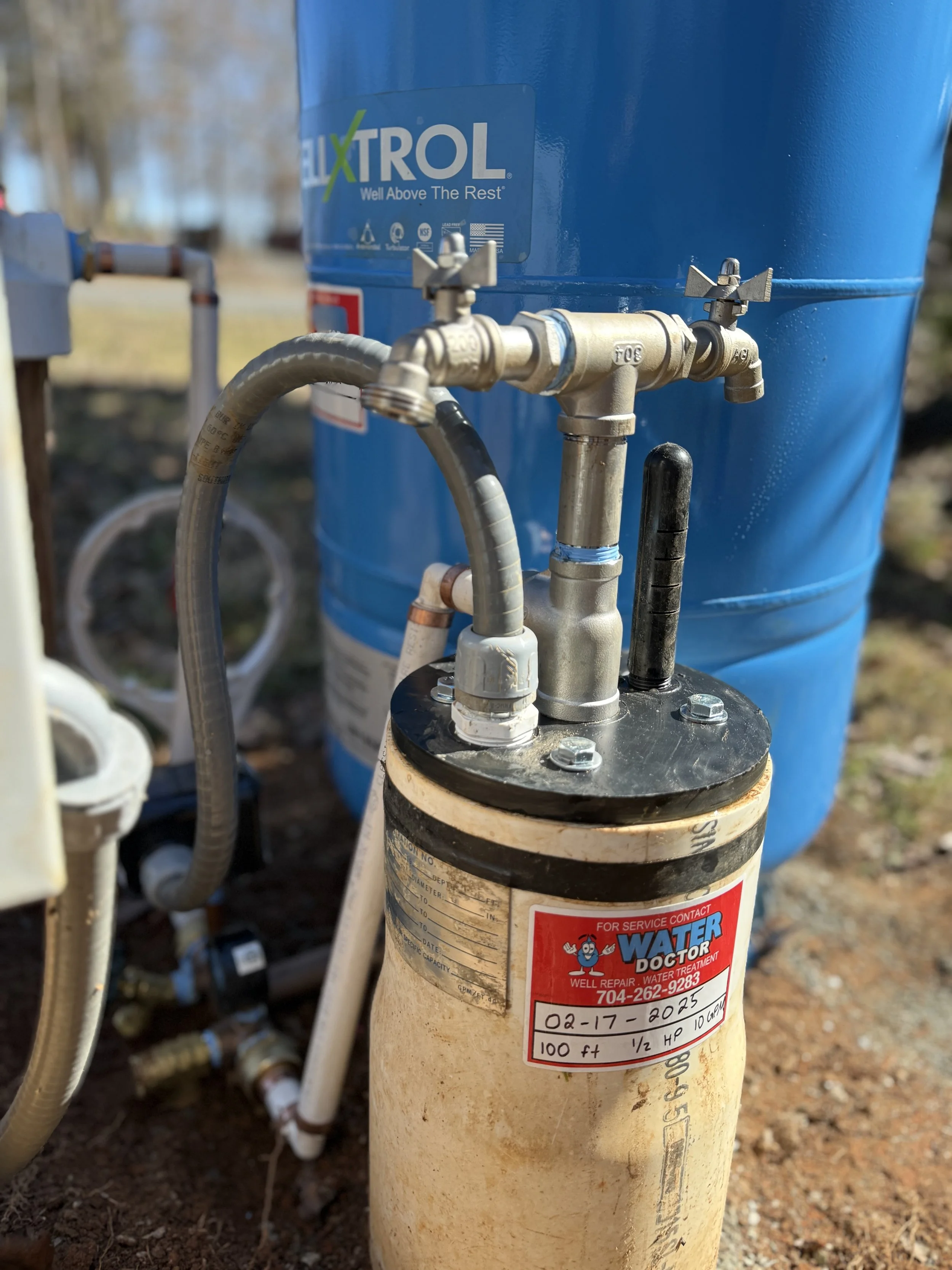 A water well system with piping, valves, and a blue filter tank in an outdoor setting.
