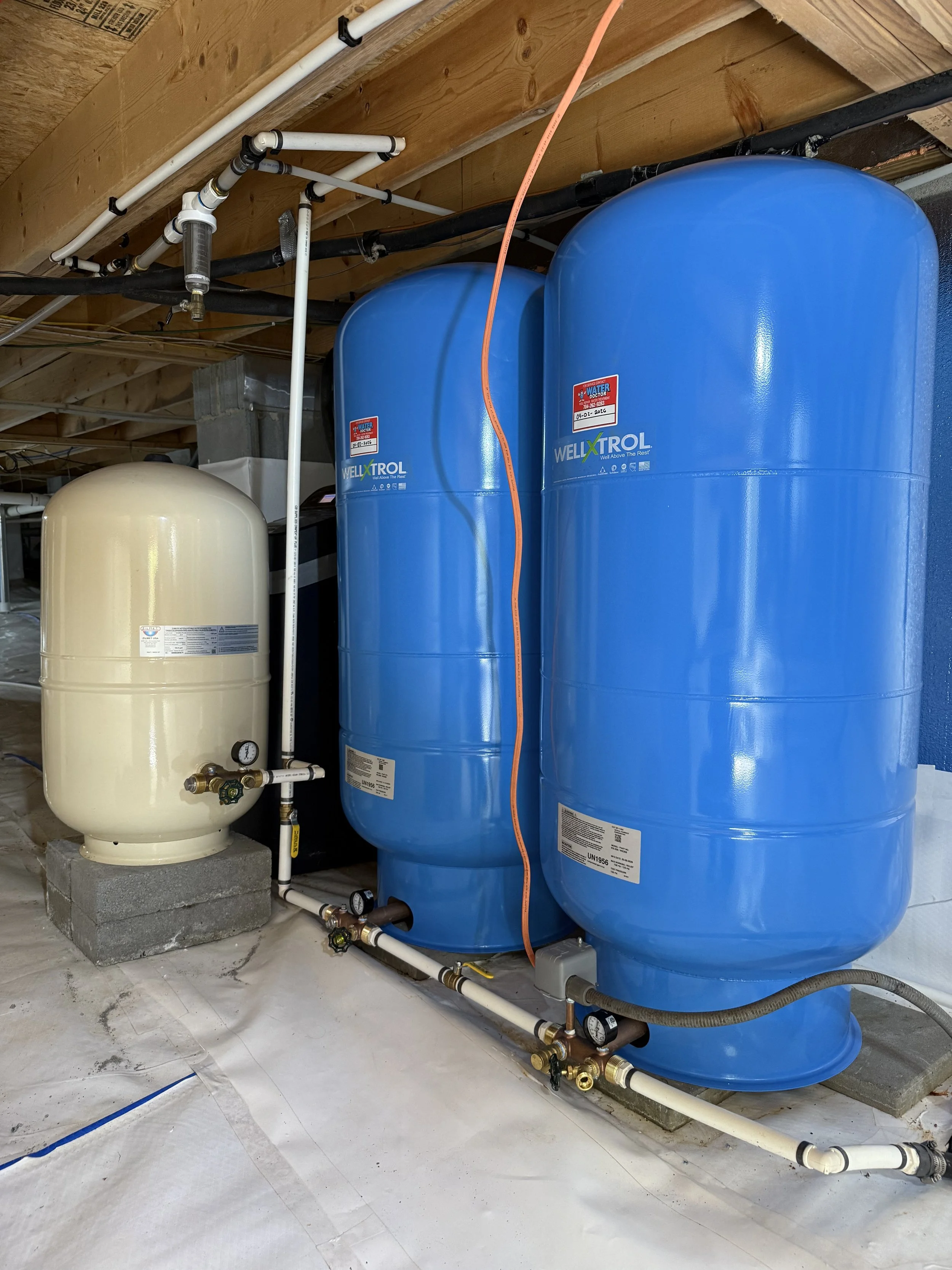 Water filtration system with a small beige tank and two large blue tanks connected by pipes and gauges, installed on a concrete floor in an attic space.