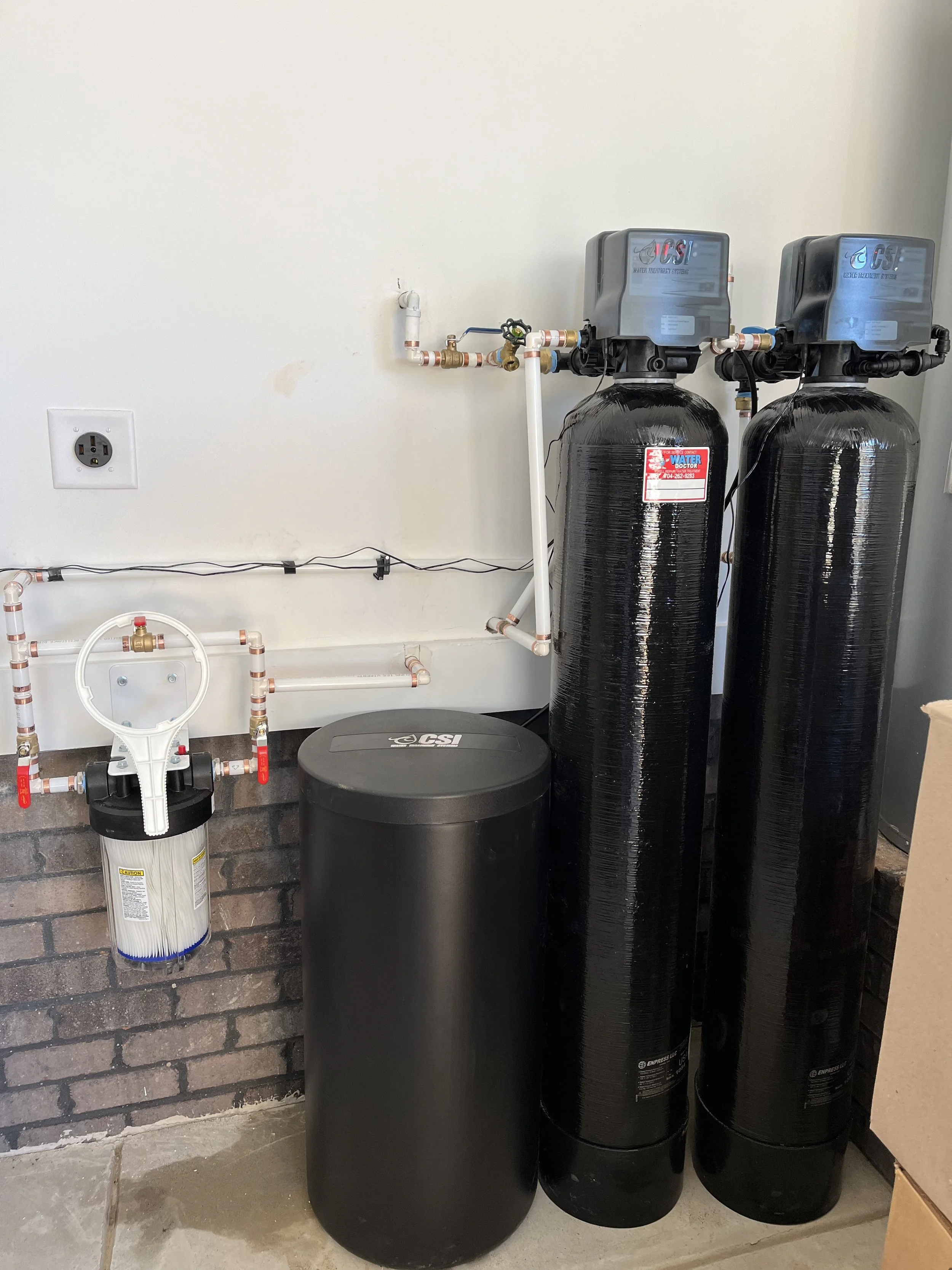 Water treatment system with two large black tanks, a smaller black cylinder, and various connected pipes and filters against a white wall.