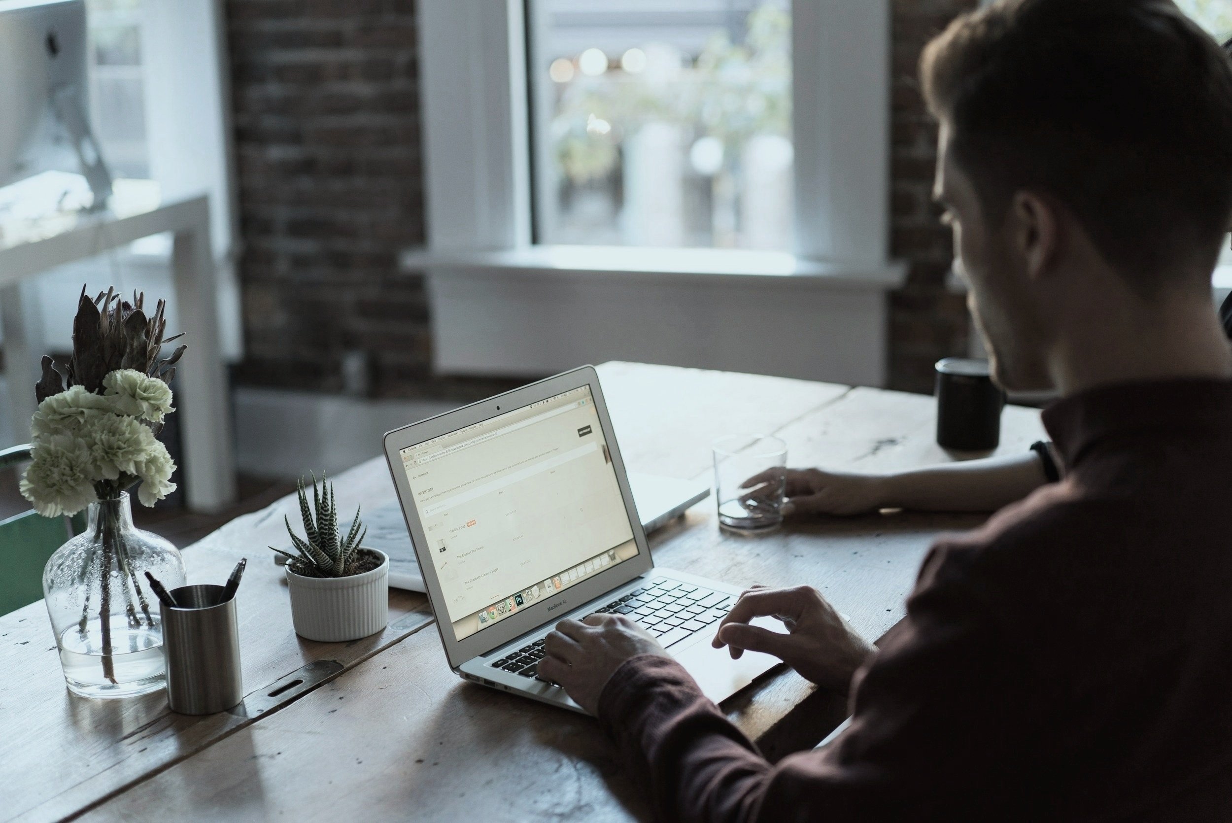 Person working on a laptop at a wooden table with a potted plant and vase of flowers, near a window with natural light.