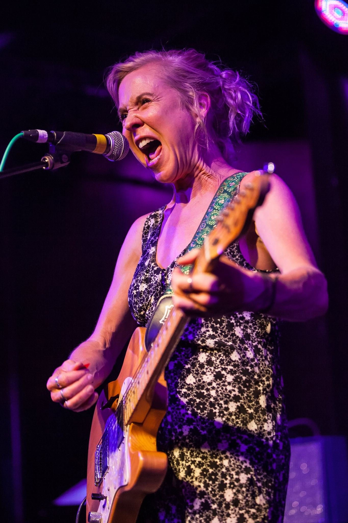Woman passionately singing and playing electric guitar on stage with a microphone, under purple lighting.