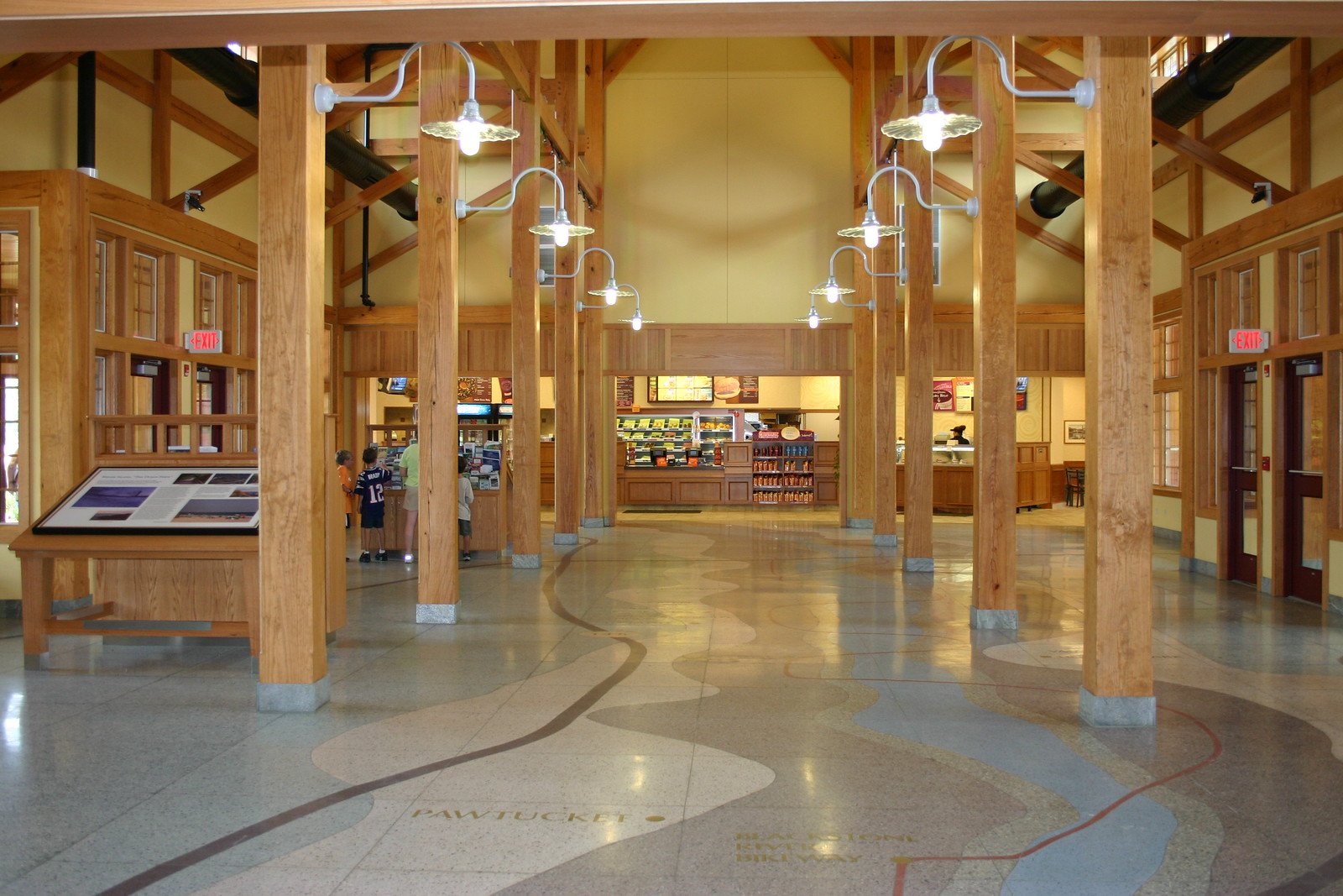 Interior of a rest area building with wooden beams, hanging lights, and informational displays. The floor features a decorative map with city names. People are visible near a counter offering snacks and books.