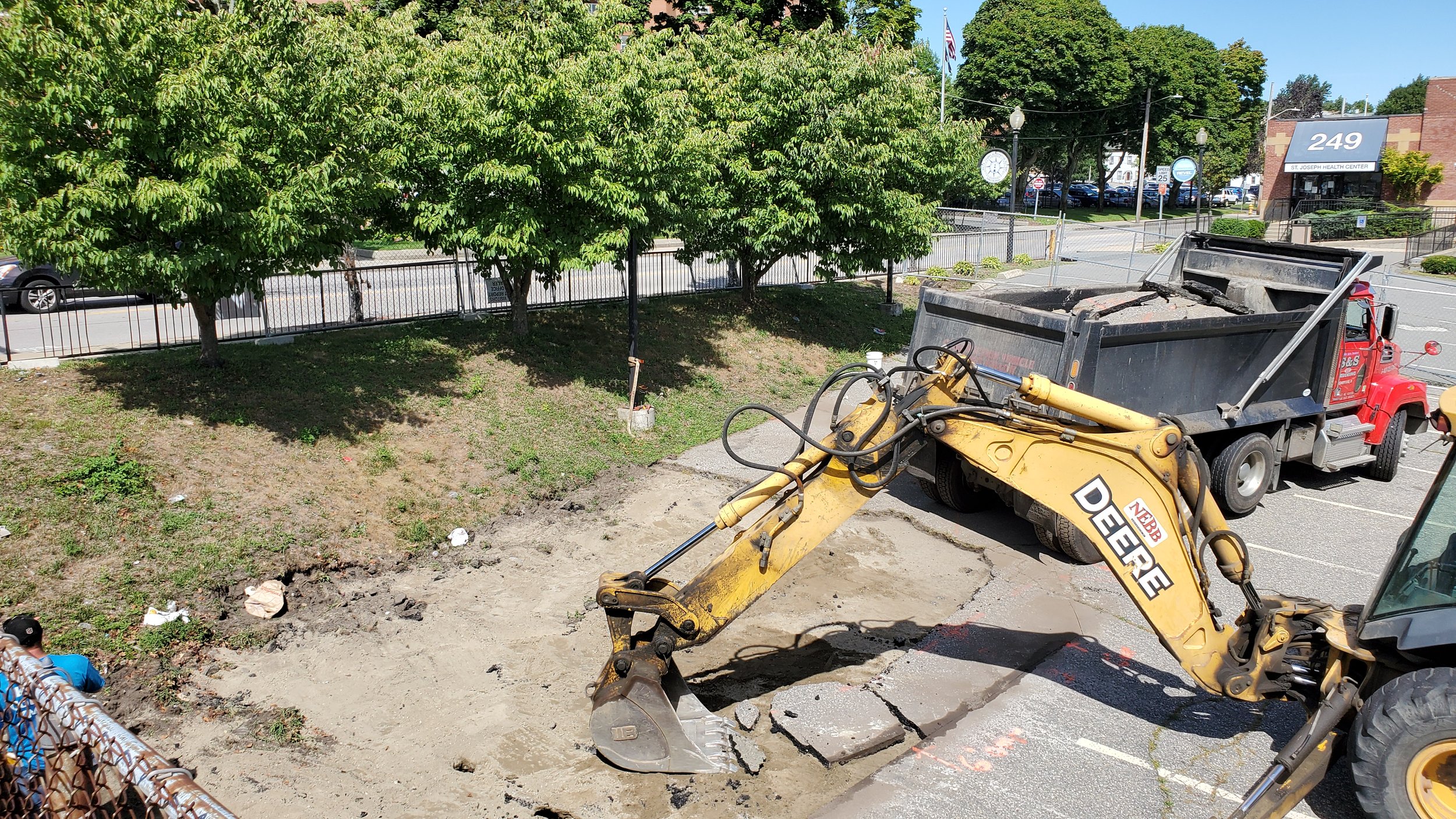 Construction site with a backhoe and dump truck, breaking up asphalt in a parking lot, surrounded by trees and a fence.