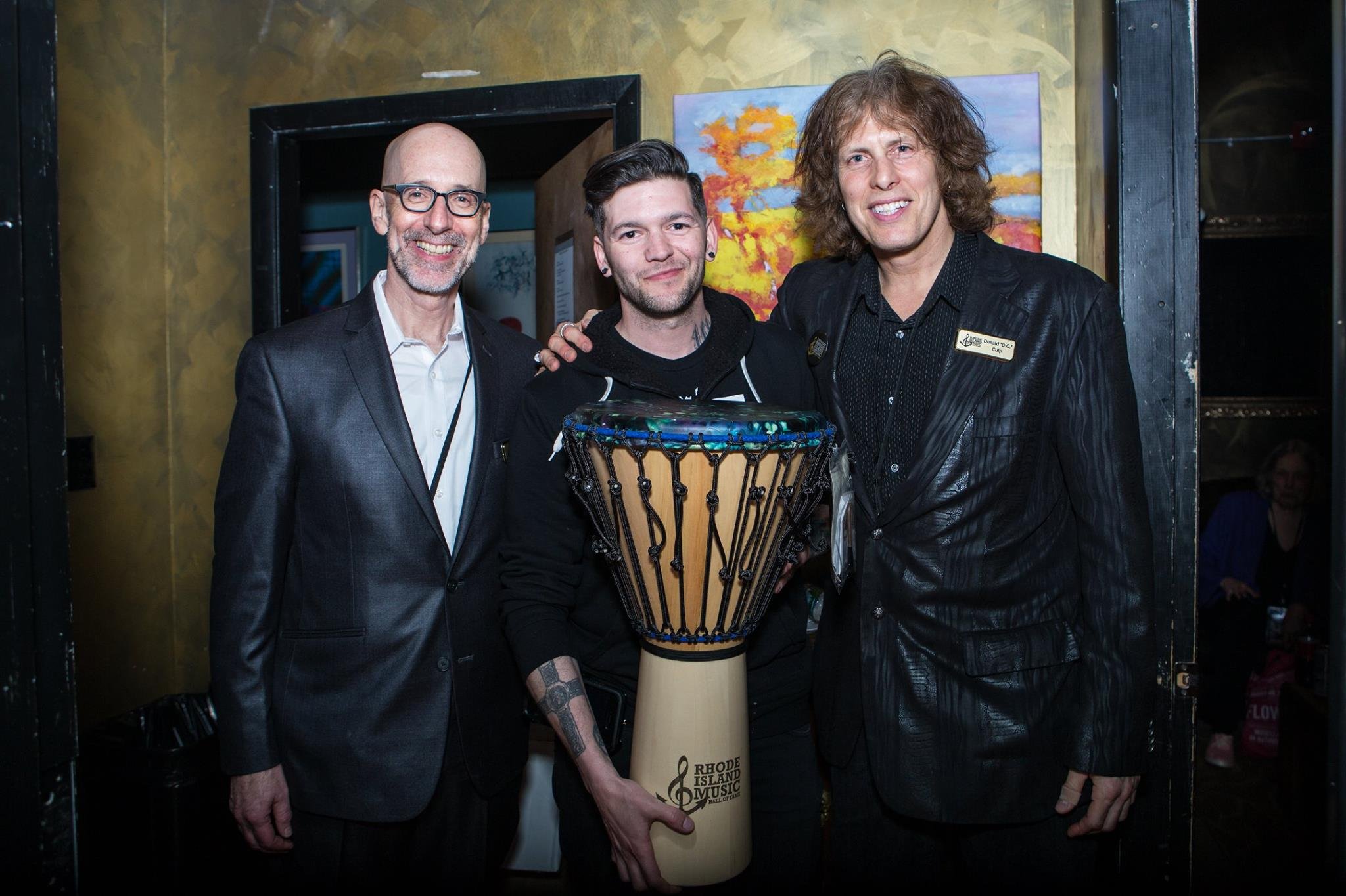 Three men smiling, standing indoors with one holding a djembe drum labeled 'Rhode Island Music.'