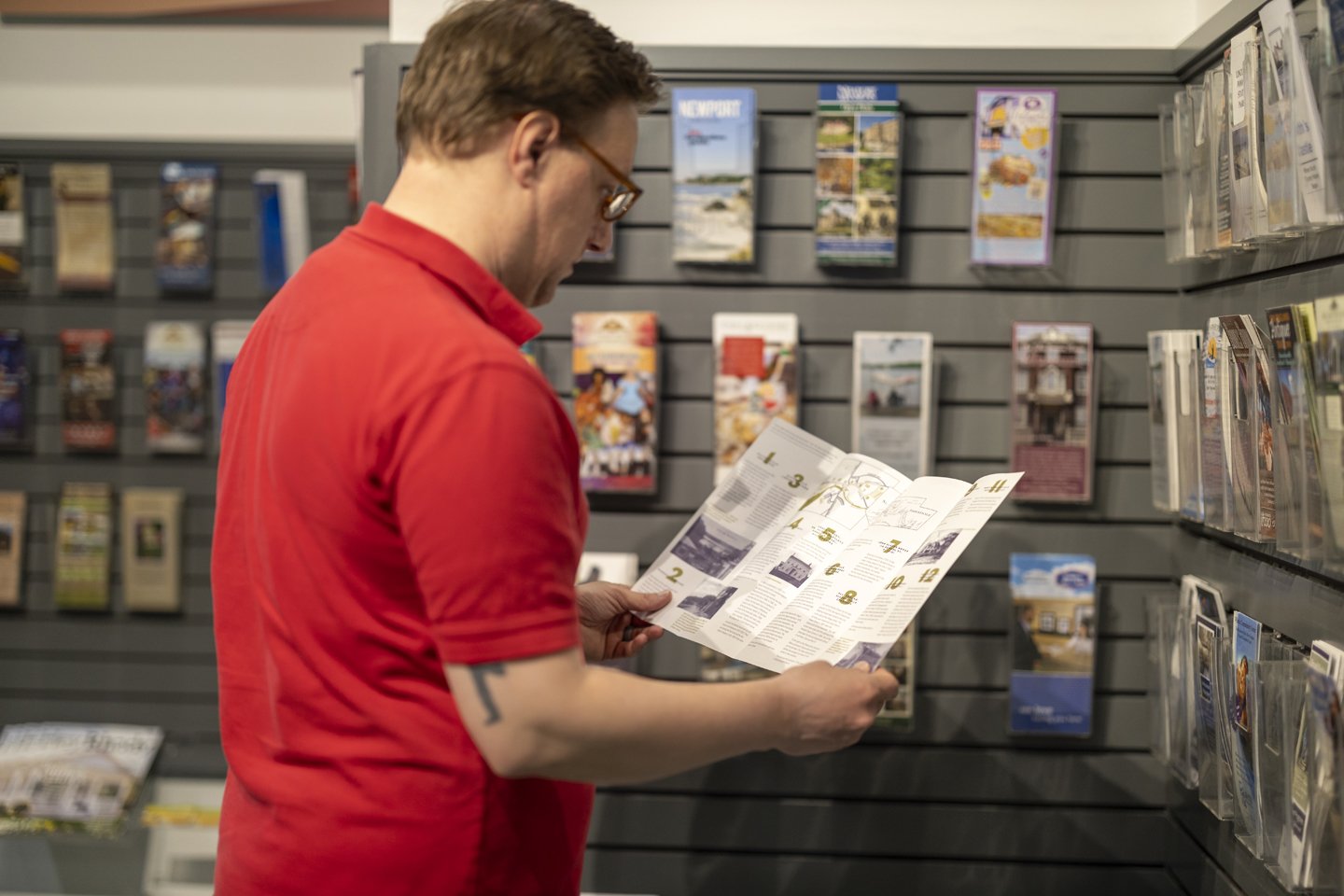 Man in red shirt reading brochure in a tourist information center with various travel pamphlets displayed on the wall.