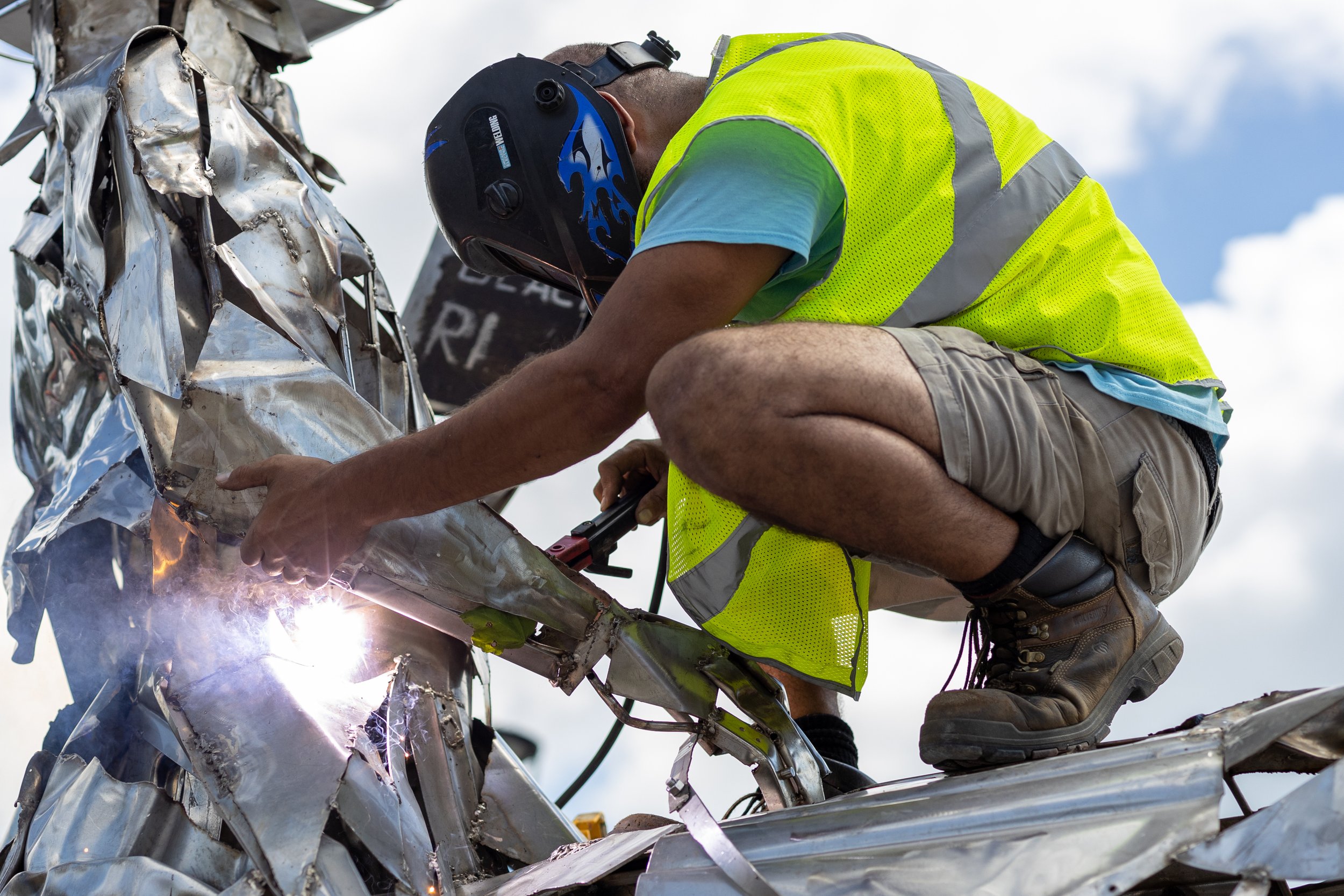 Person welding metallic sculpture outdoors wearing safety gear