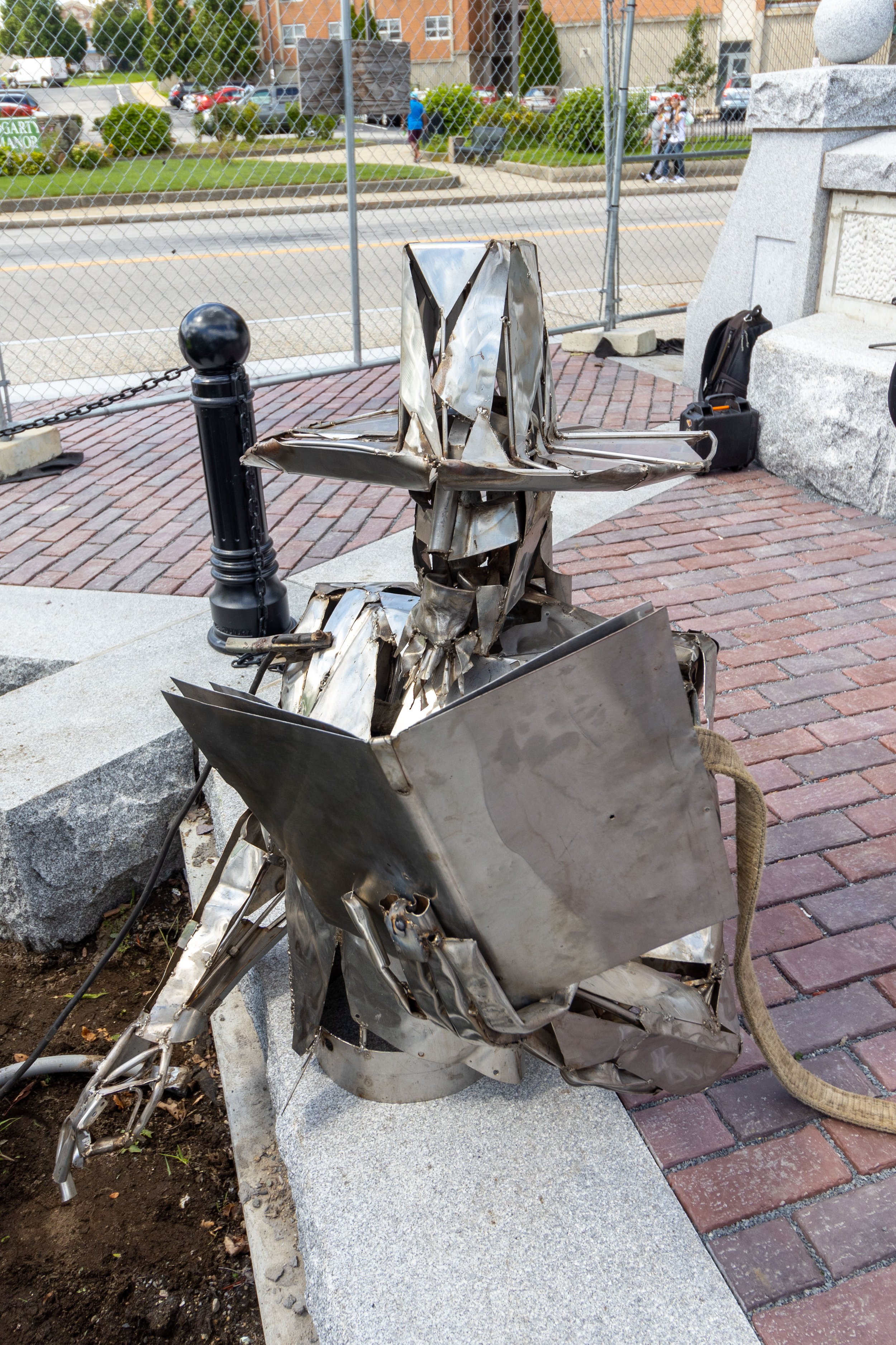 Metal sculpture of a figure reading a book, wearing a wide-brimmed hat, situated on a brick walkway.