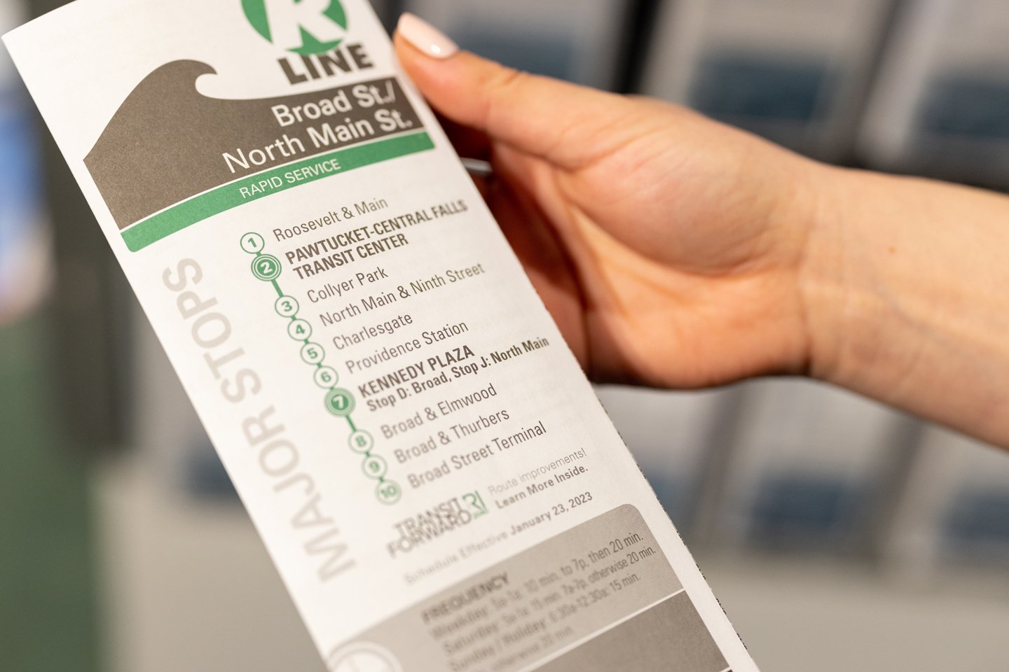 A person holding a public transportation brochure for the "R Line," detailing major stops such as Broad St, North Main St, and others. The schedule is effective January 23, 2023, and includes information on rapid service and route improvement details