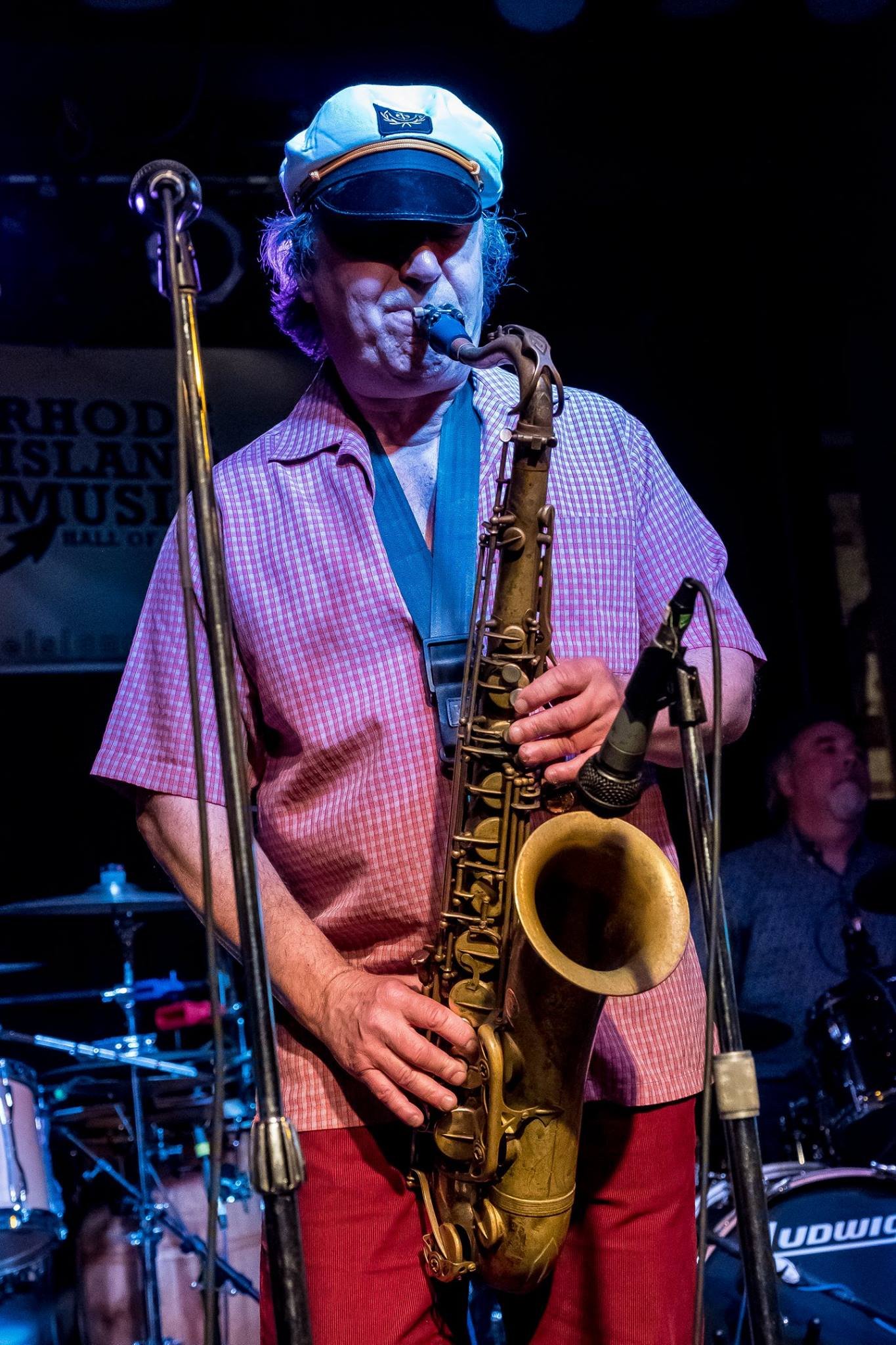 A man wearing a captain's hat plays the saxophone on stage, with a microphone and drum set in the background.
