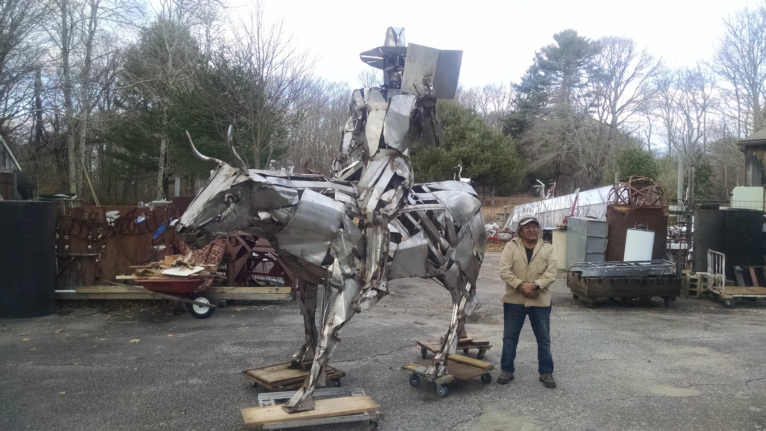 Metal sculpture of a cowboy on a horse next to a person standing, surrounded by trees and miscellaneous objects.