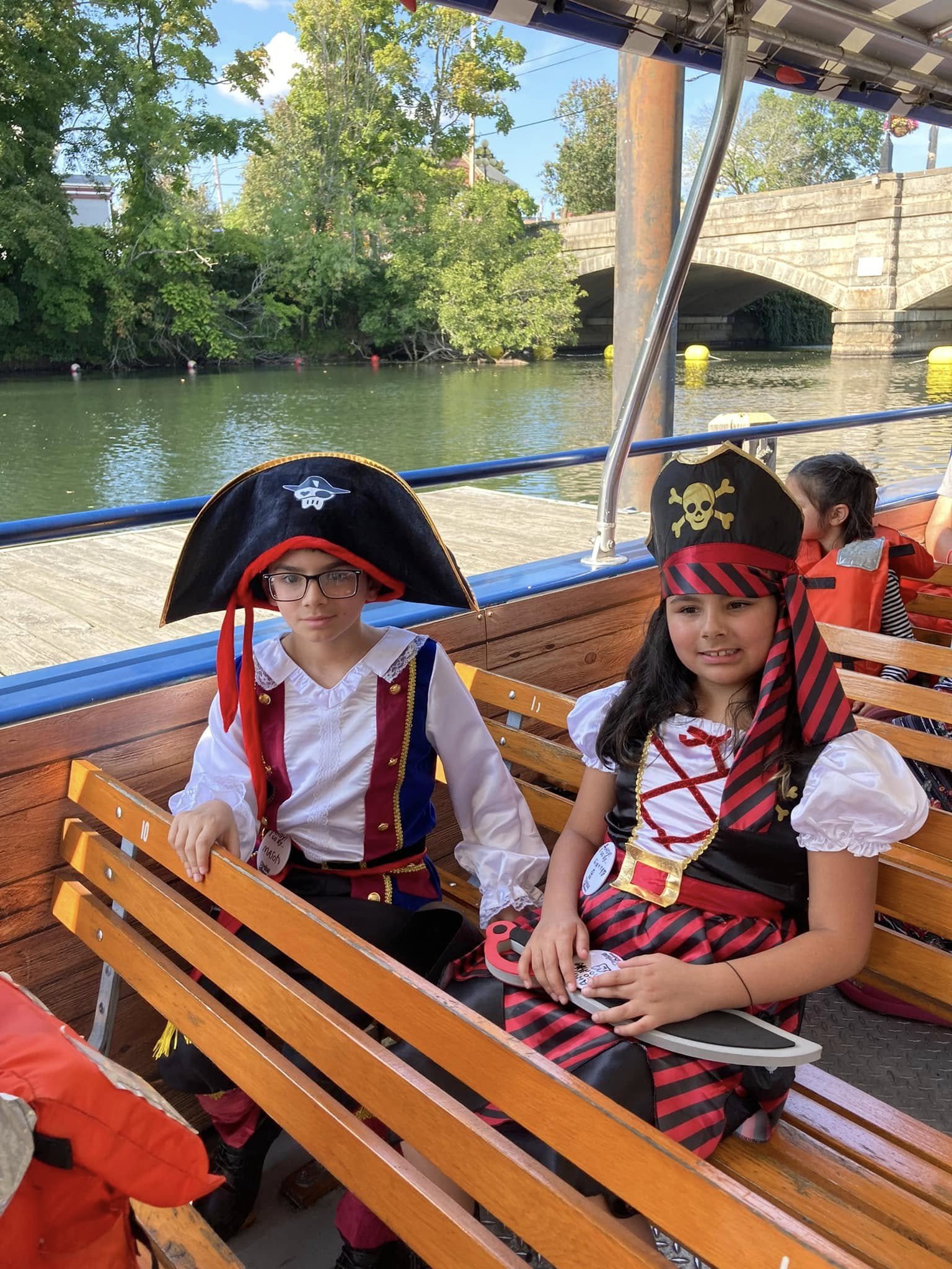 Two children dressed in pirate costumes sitting on a wooden bench of a boat on a river. The background includes trees, a bridge, and a clear sky.
