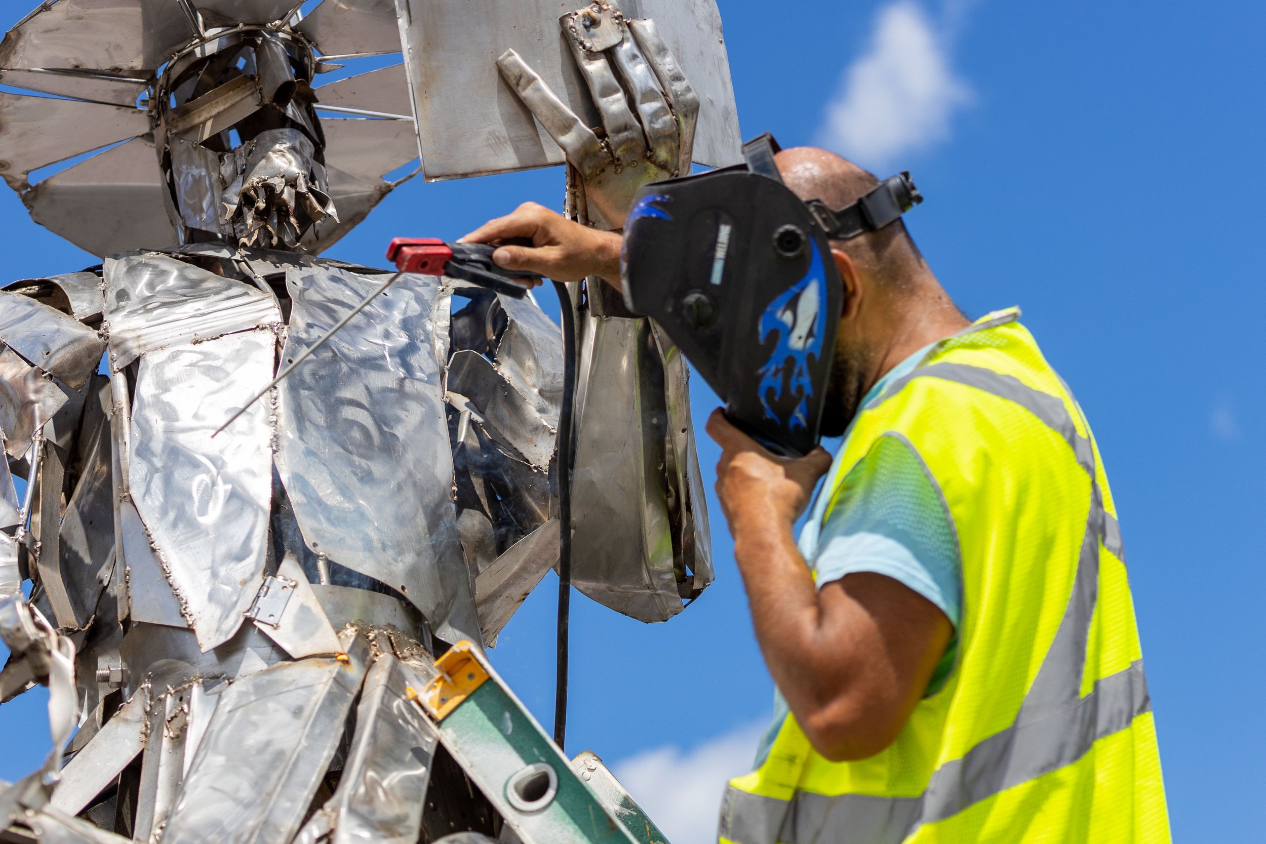 Person welding a metal sculpture outdoors, wearing a safety vest and welding helmet.