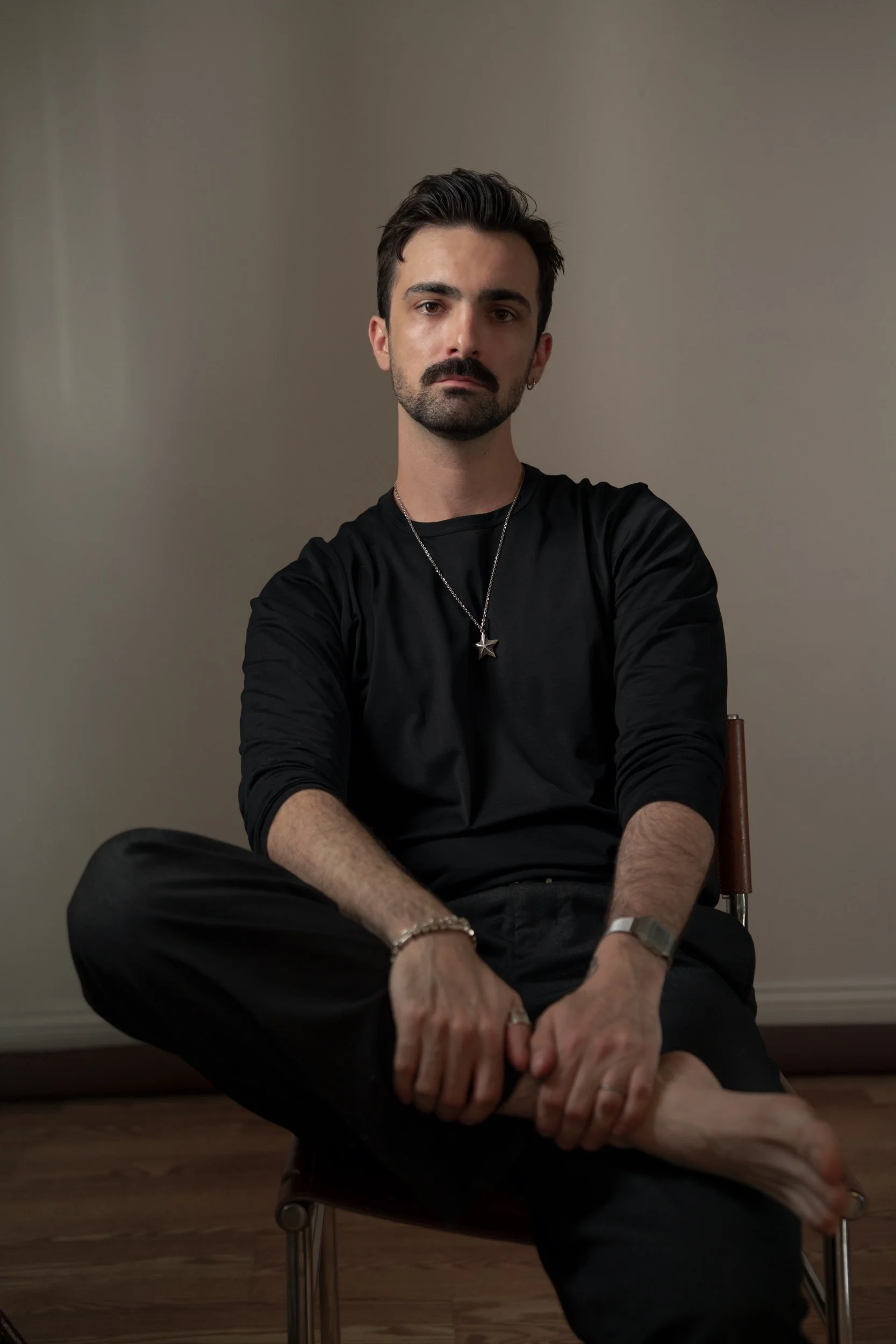 A young man with dark hair, a beard, and a mustache, sitting on a wooden chair with a neutral expression, wearing a black long-sleeved shirt, black pants, a star necklace, wrist accessories, and holding his leg with his foot resting on the chair's edge.