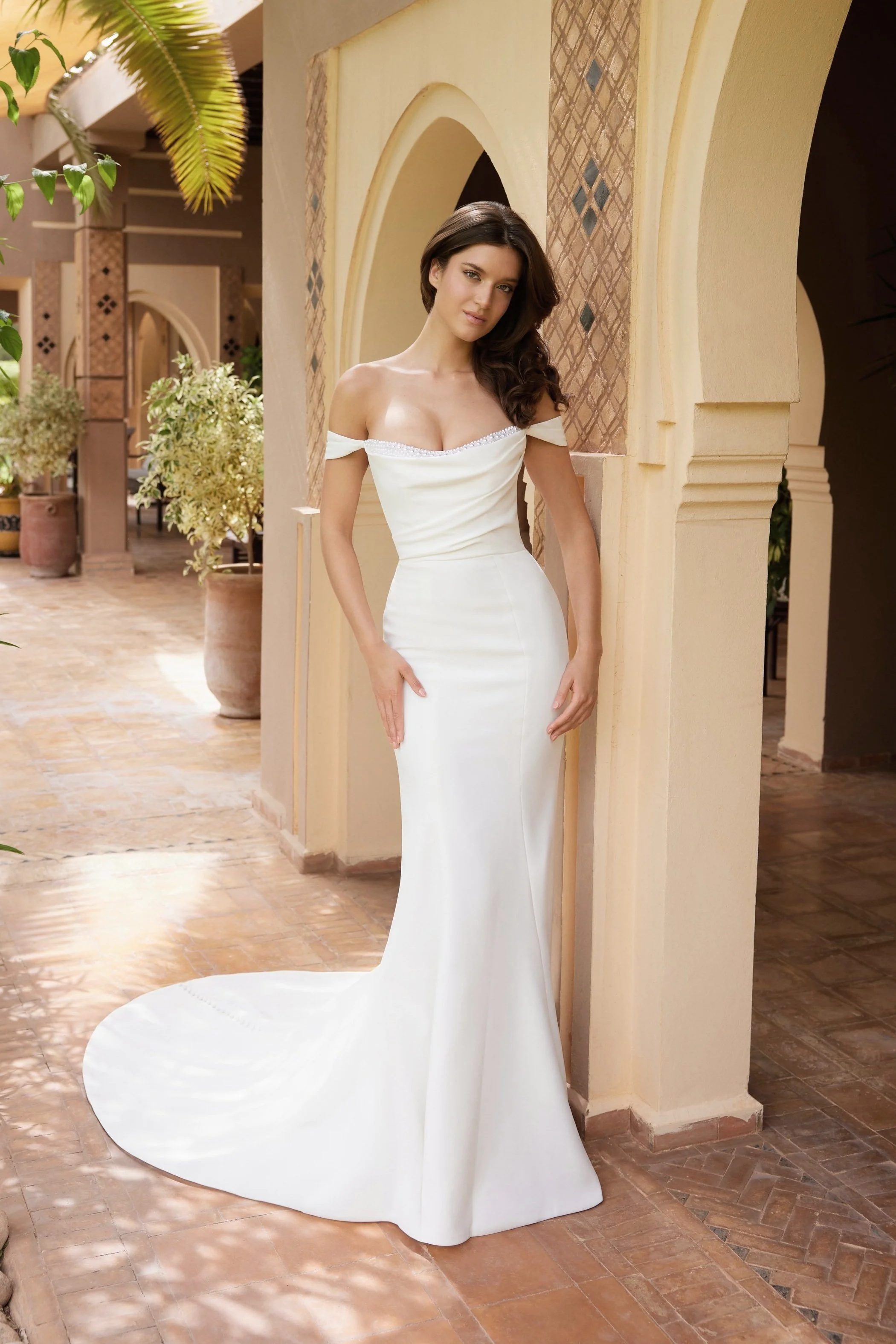 A woman in a white off-the-shoulder wedding dress with a train, standing in a sunny, Mediterranean-style courtyard with potted plants and arched doorways.