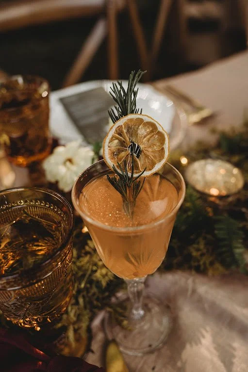 A cocktail in a crystal glass with sprig of rosemary and slice of dried lemon on a beautifully set table.