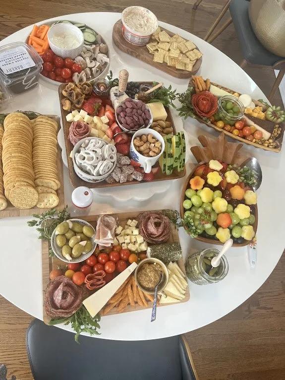 A glass of red beverage with orange slices on a dark table surrounded by a cheese and charcuterie plate, olives, and a plate of cookies.