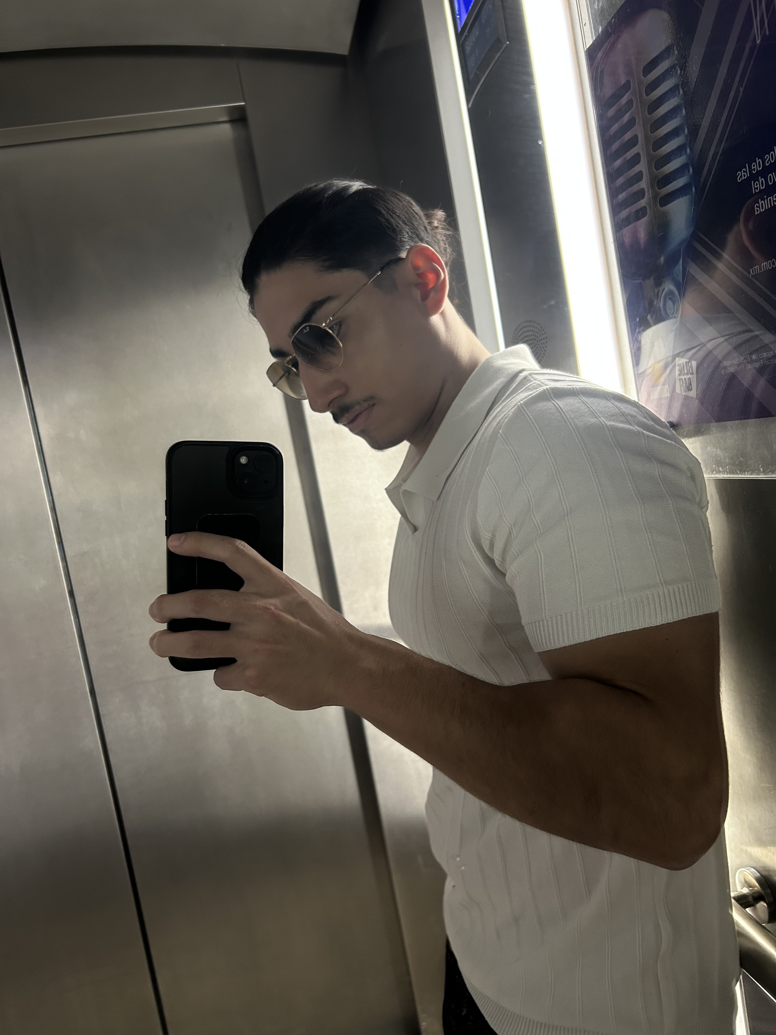A man taking a mirror selfie in an elevator, wearing sunglasses and a white shirt with vertical pattern.