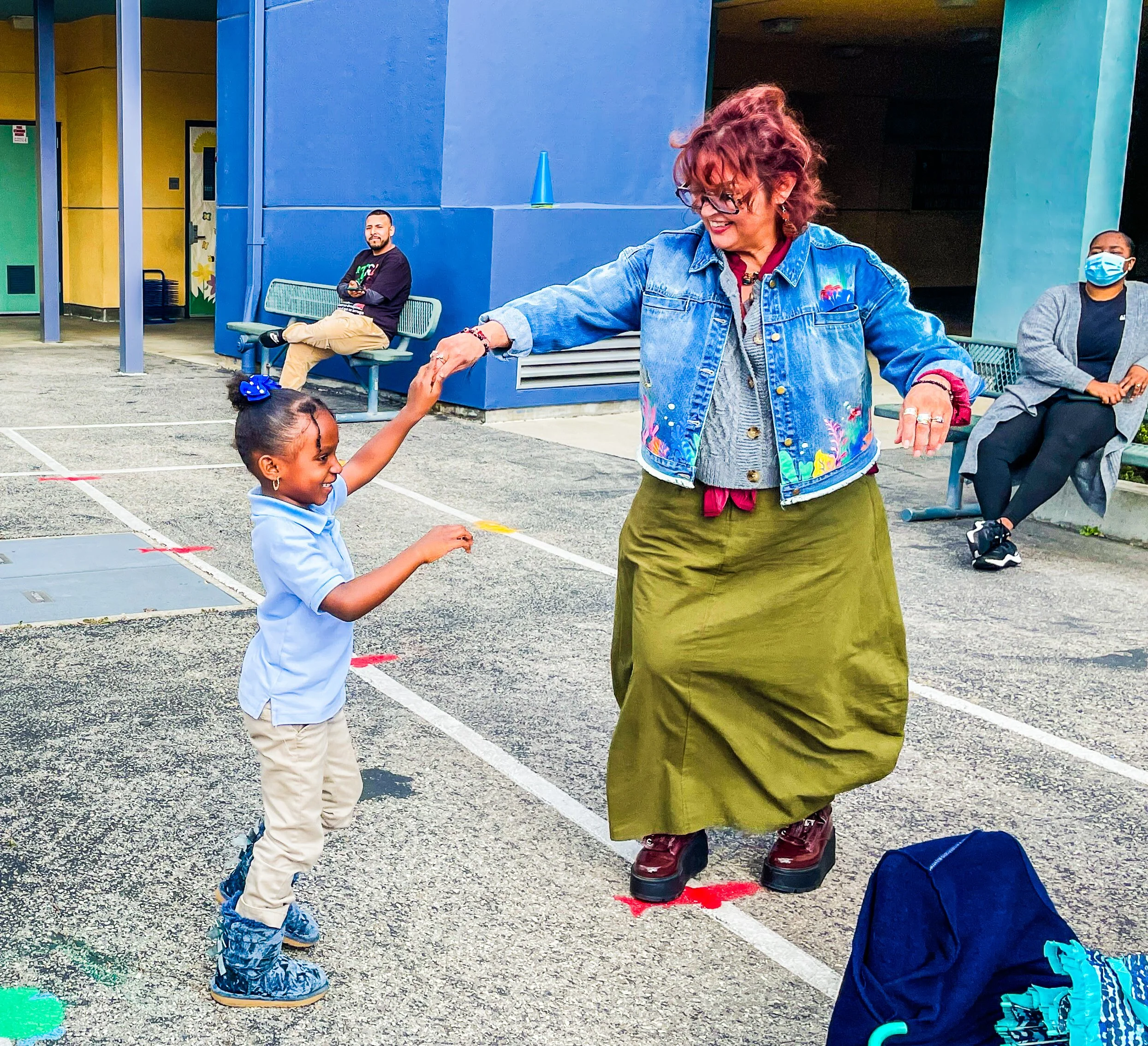 A young girl dancing with an older woman on a marked outdoor ground, while two people are sitting on benches in the background, one wearing a face mask and the other holding a cellphone.
