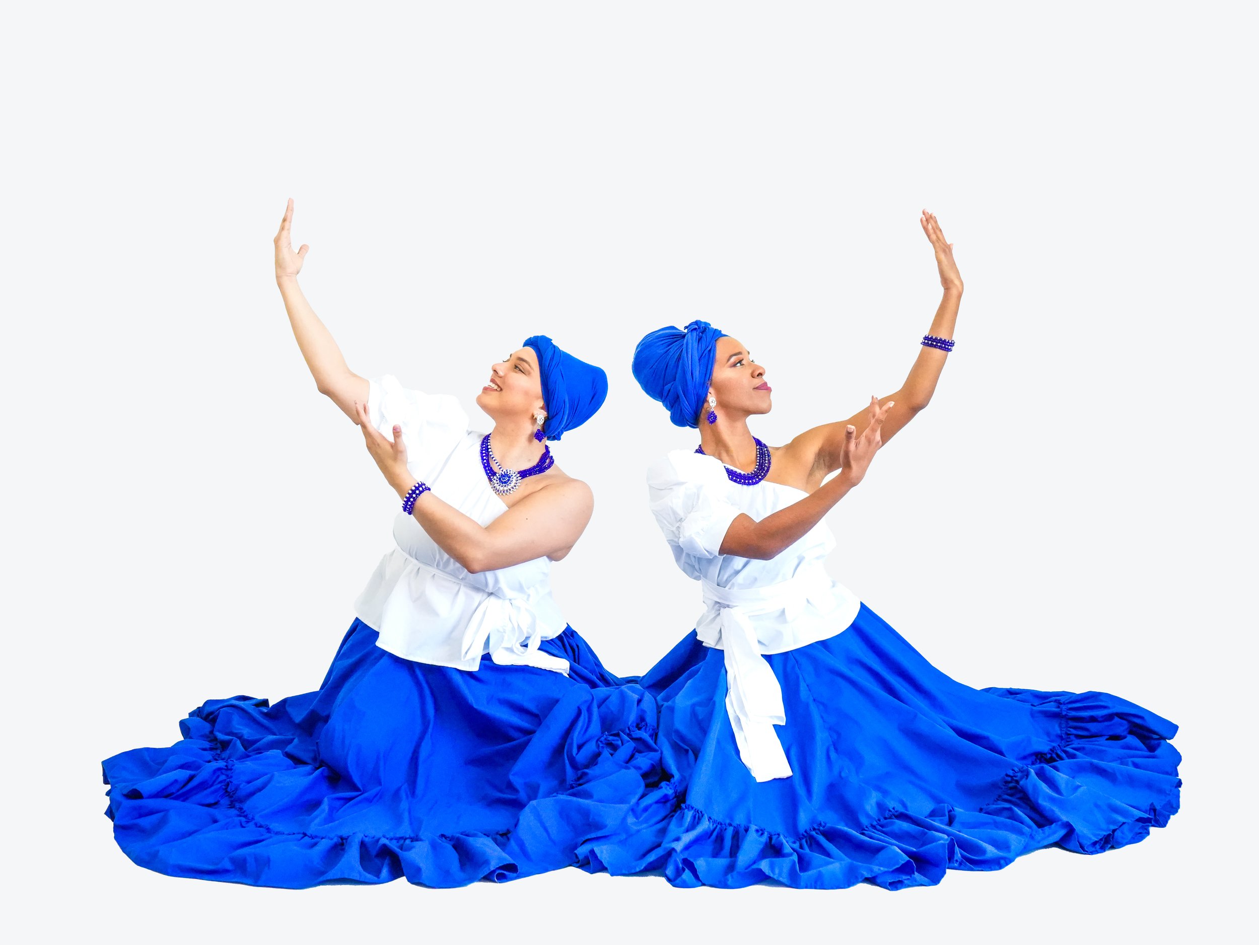 Two women dressed in traditional blue and white dance costumes, sitting on the floor with long, ruffled blue skirts, and wearing matching blue headscarves, jewelry, and white tops, striking graceful poses against a plain white background.