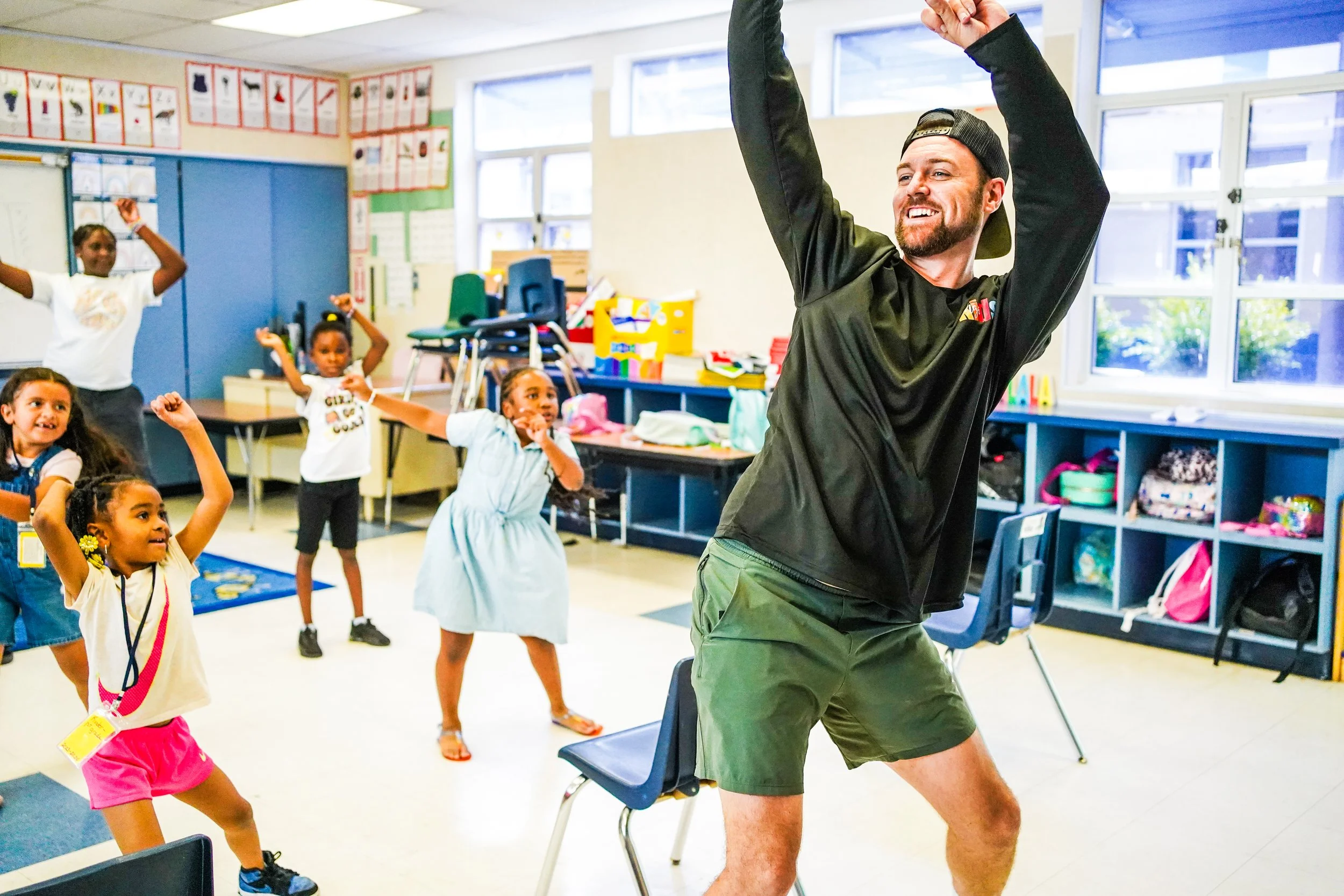 A man leading a dance or exercise class for a group of young children in a classroom, with all participants smiling and raising their arms.