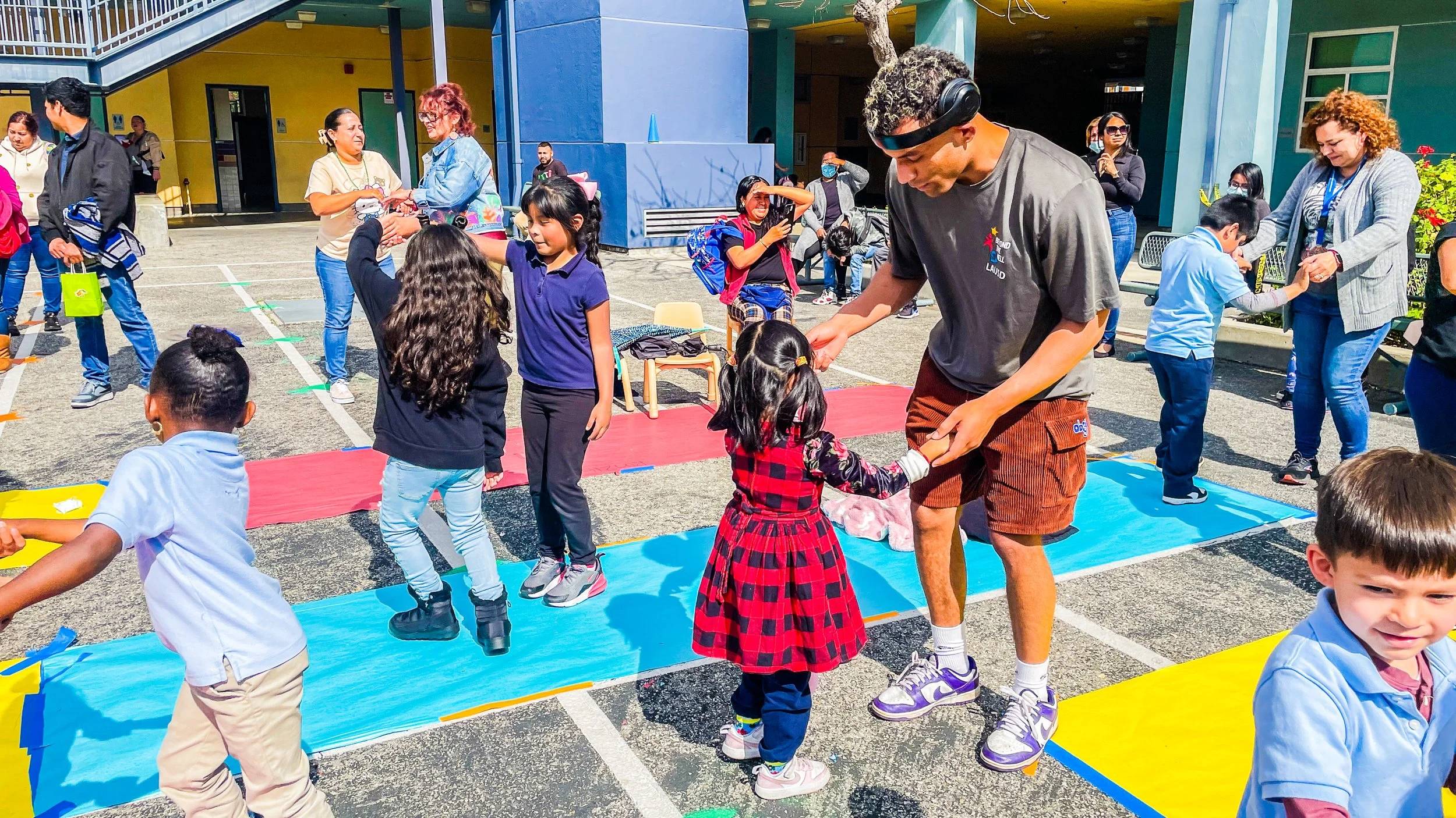 Children and adults dancing and playing on colorful mats outdoors at a school event.
