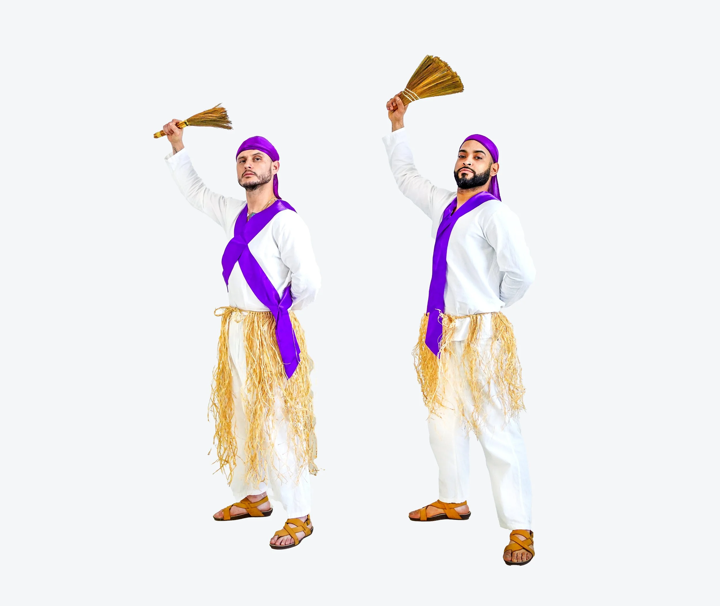 Two men dressed in white outfits with purple sashes and headbands, holding brooms and wearing straw skirts against a plain background.