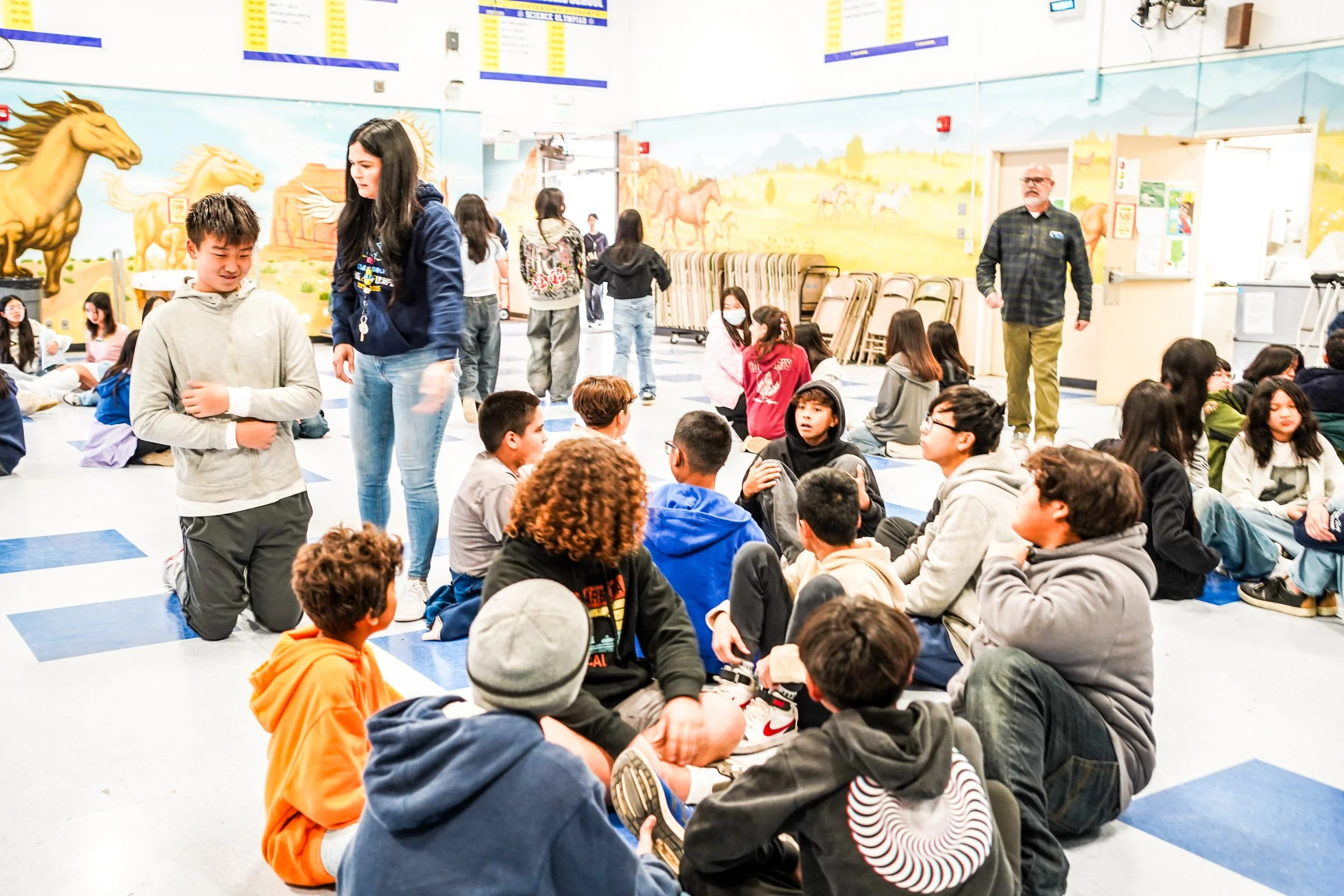 A group of children and teenagers sitting and talking in a colorful classroom with mural paintings of horses and mountains on the walls.