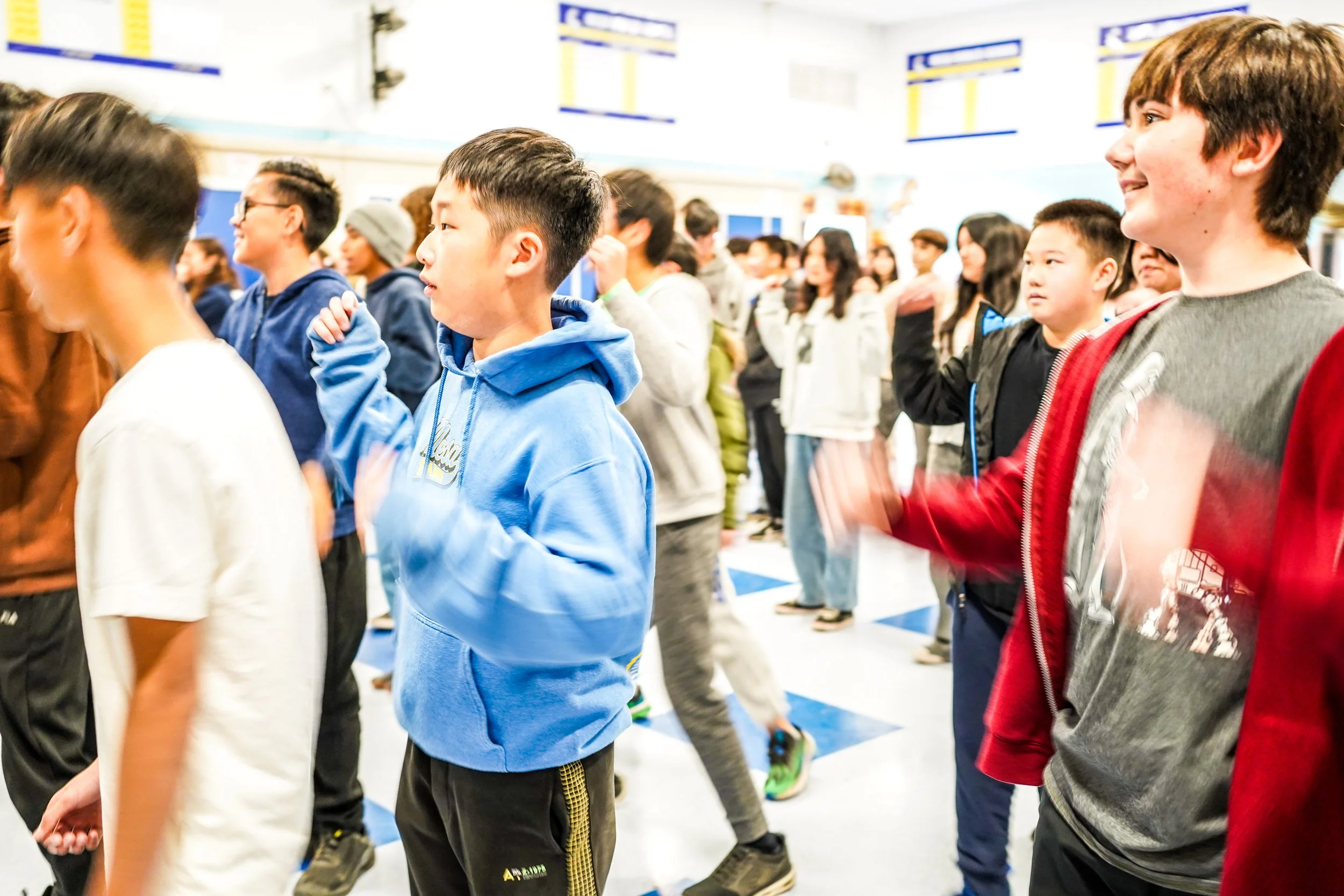 A group of children and teenagers standing in a gymnasium, participating in a group activity or dance, with some raising their hands and others moving their bodies, all facing the same direction.