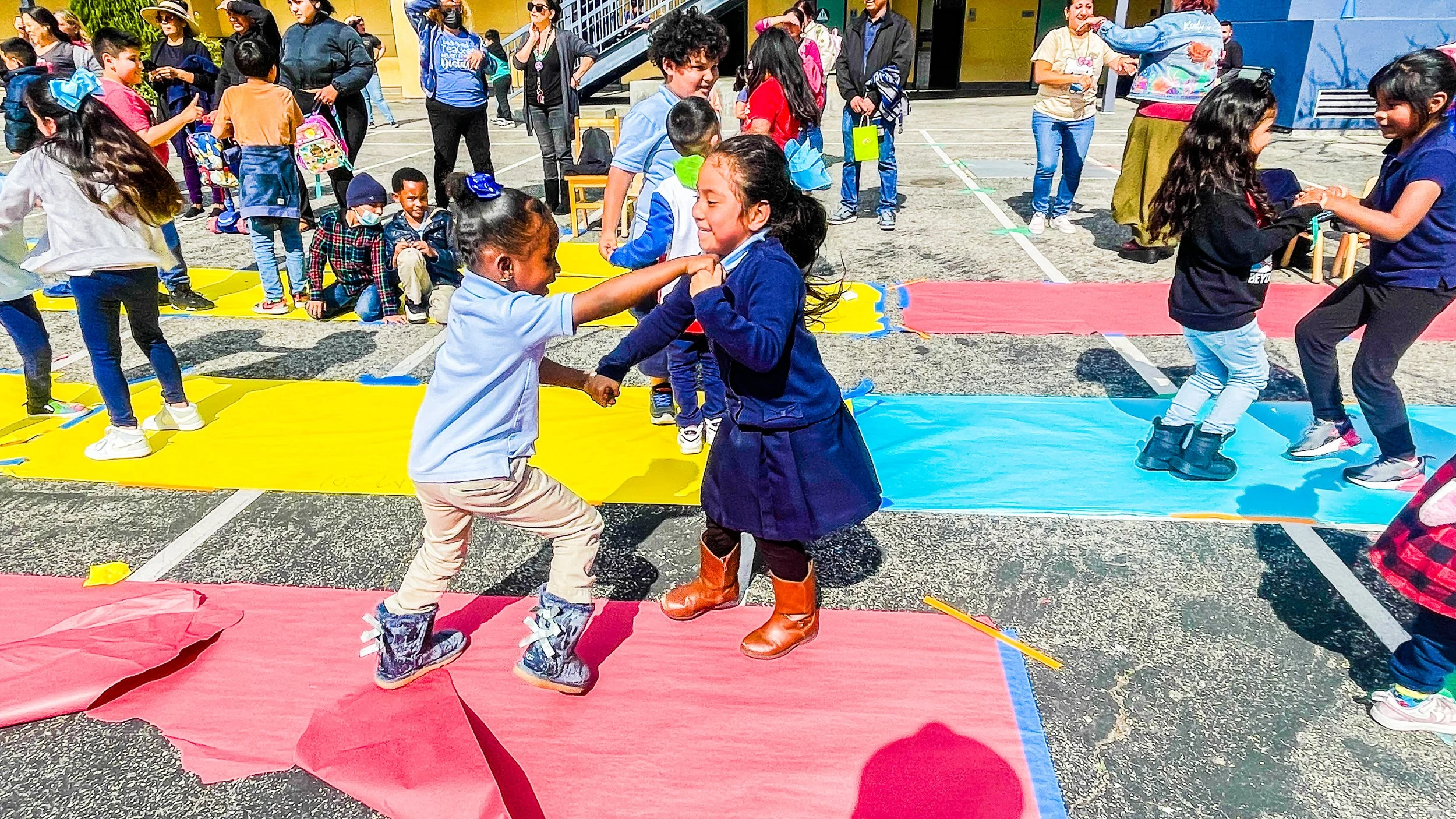 Children playing and dancing on colorful mats outdoors at a school event with teachers and parents in background.