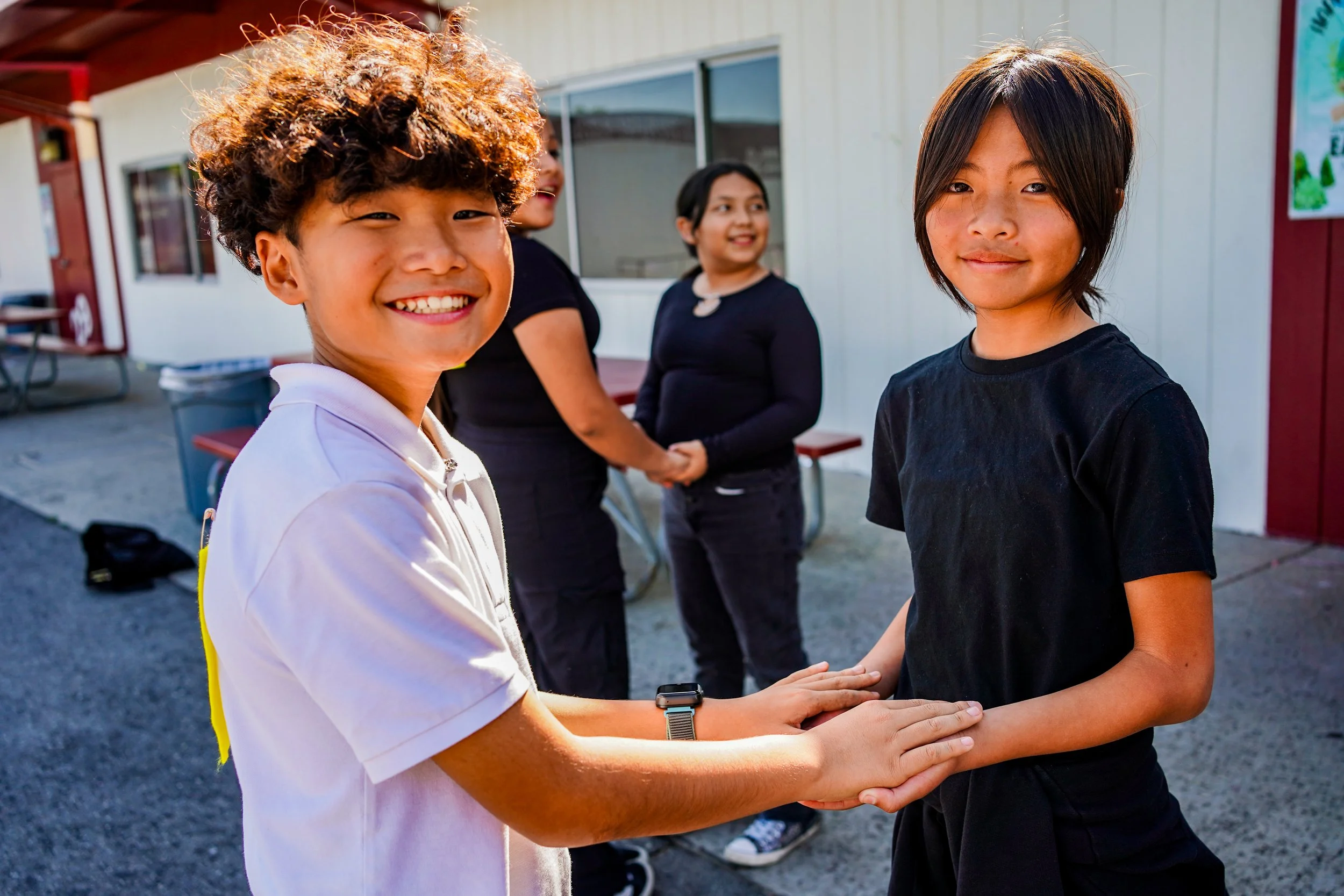 Two boys handshake smiling in the foreground, with two girls smiling in the background outside a building.