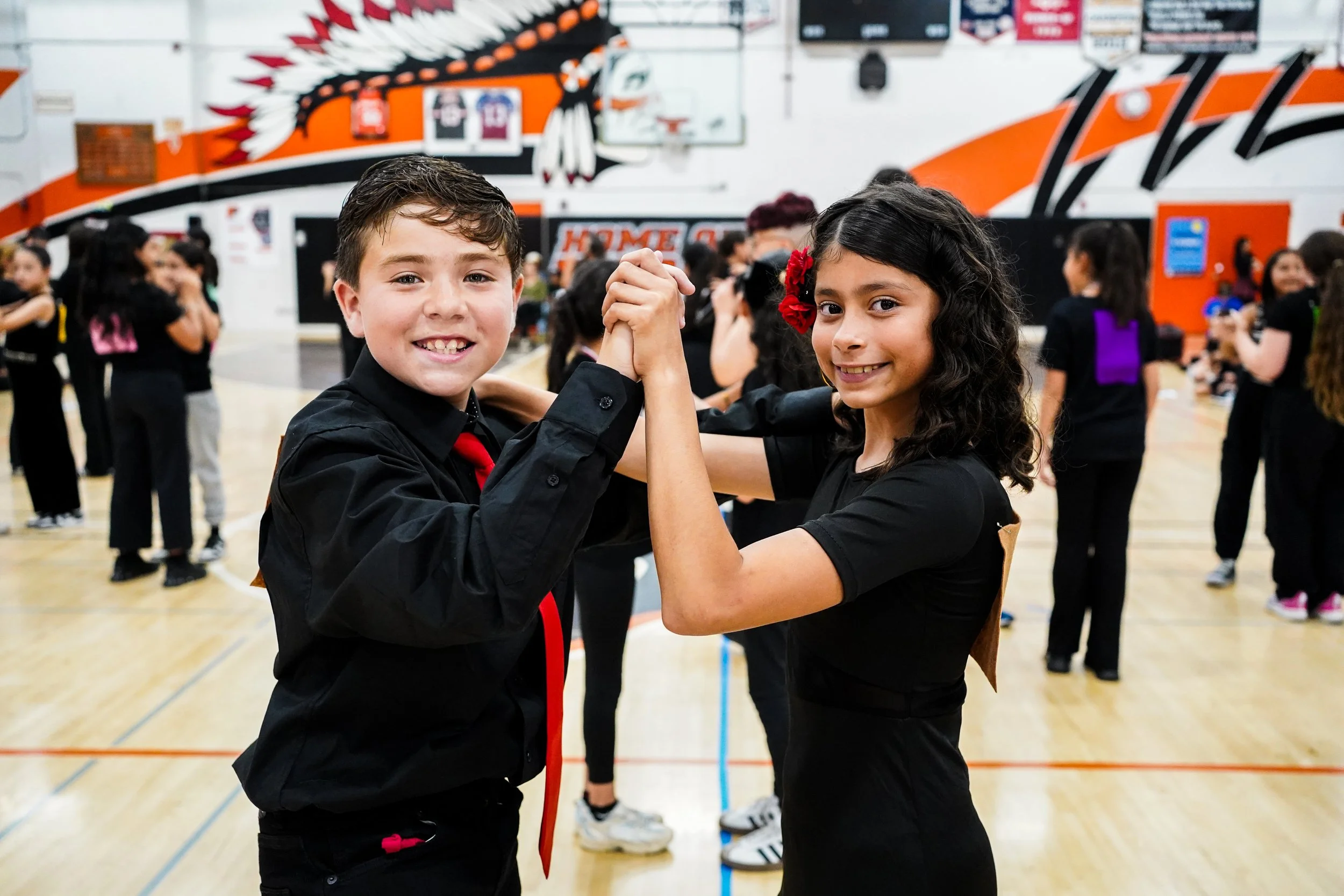 A young boy and girl are smiling and holding hands in an indoor gymnasium, surrounded by other children, during a school dance or event.