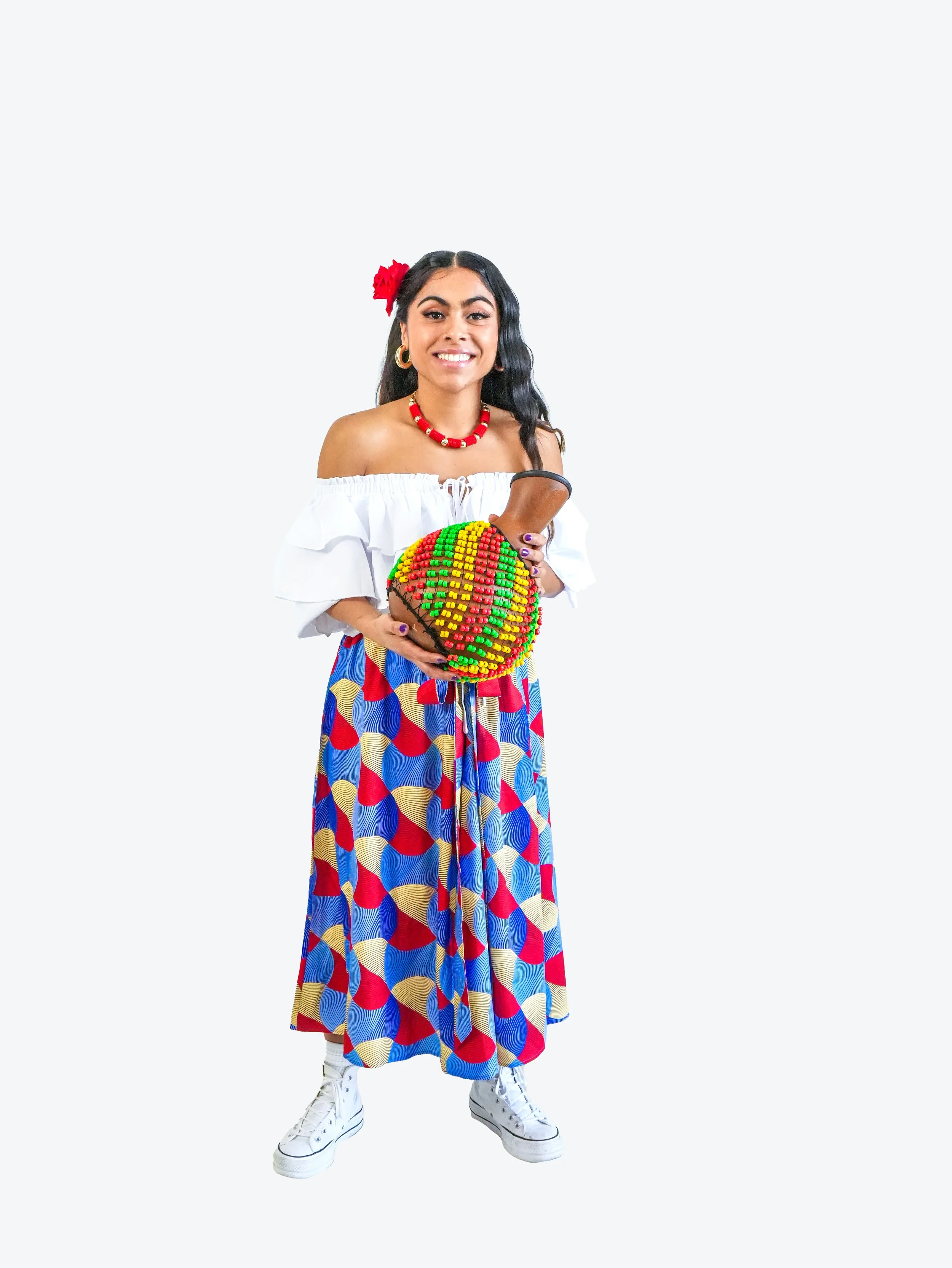 Young woman in a white off-shoulder top, colorful patterned skirt, white sneakers, holding a decorated rattan ball, with a red flower in her hair, standing against a plain background.