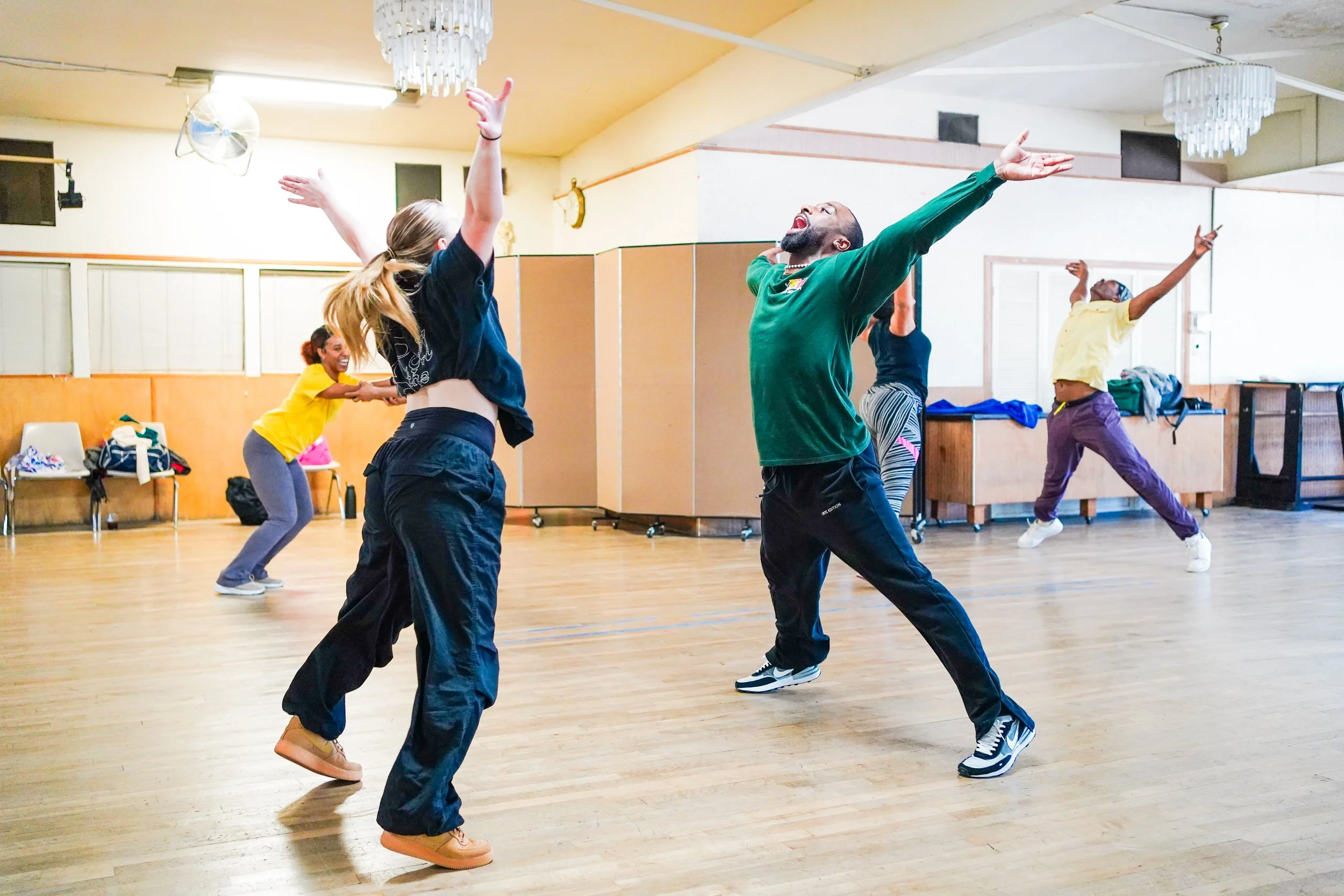 People dancing and having fun in a dance studio with wooden floors, chandeliers, and a mirror. They are mid-move with arms raised, smiling and energetic.