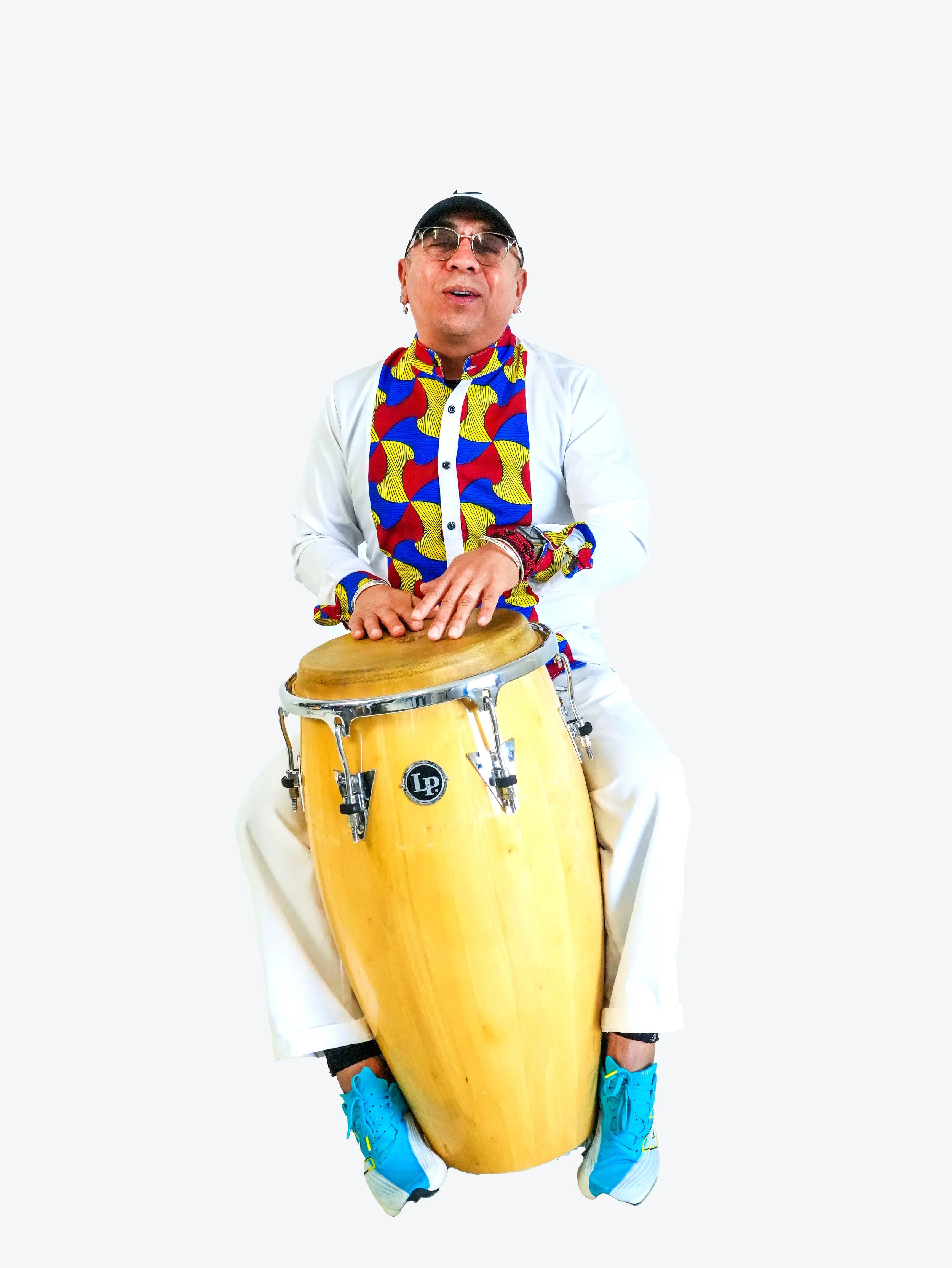 Man wearing sunglasses, baseball cap, colorful shirt, white pants and blue shoes, sitting and playing on a large conga drum.