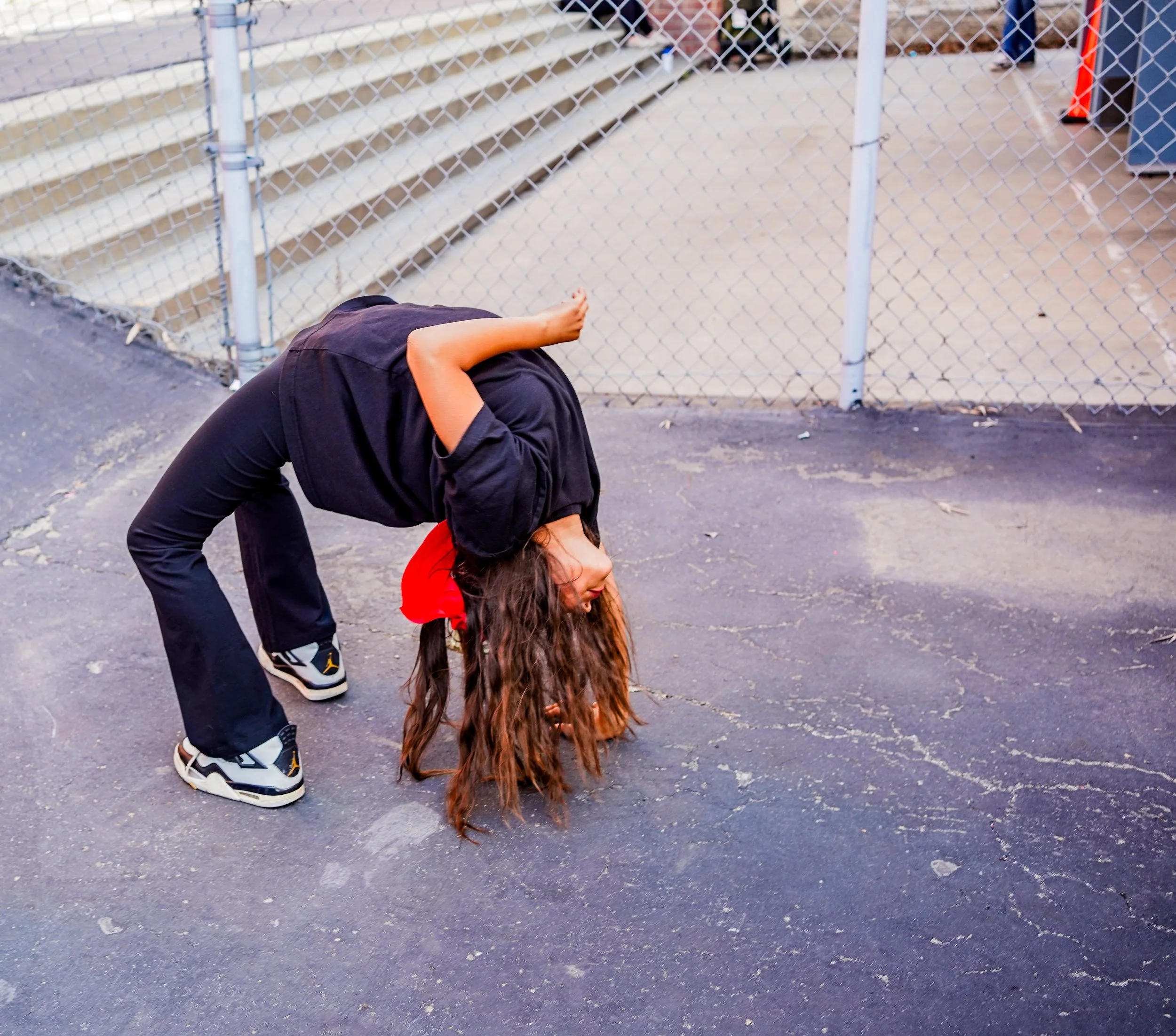 A person with long, curly brown hair bends backward with hands touching the ground. They are wearing a black hoodie, black pants, and sneakers, standing on a paved surface near a chain-link fence with stairs behind them.