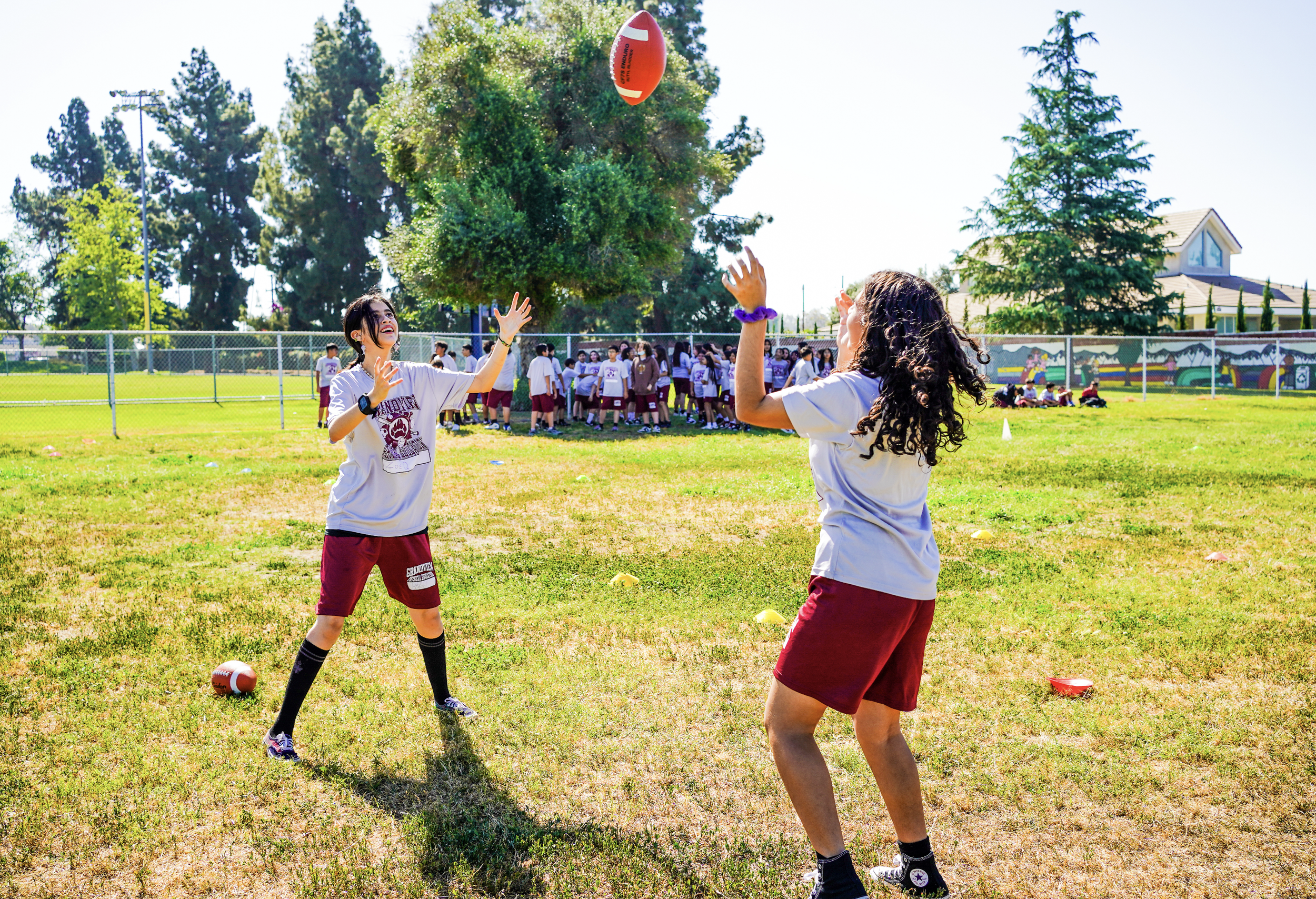 Two girls playing volleyball outdoors on a grassy field during daytime with a group of students in the background.