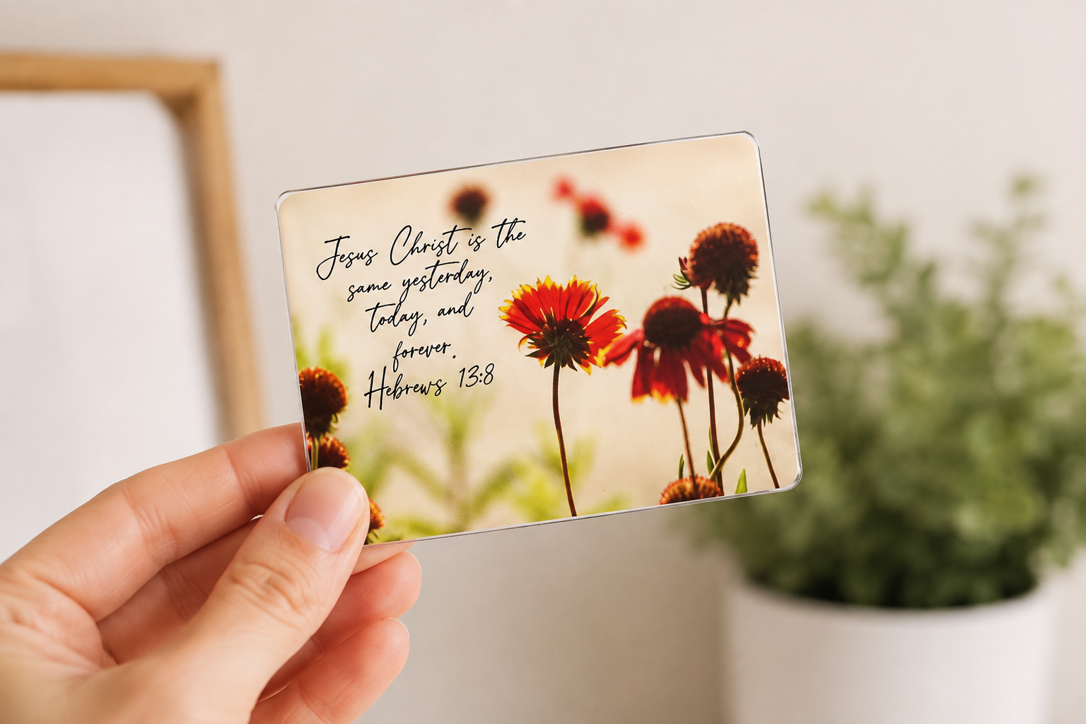 A hand holding a transparent card with a quote from Hebrews 13:8, featuring a background of red and yellow flowers, with a blurred green plant in a white pot in the background.
